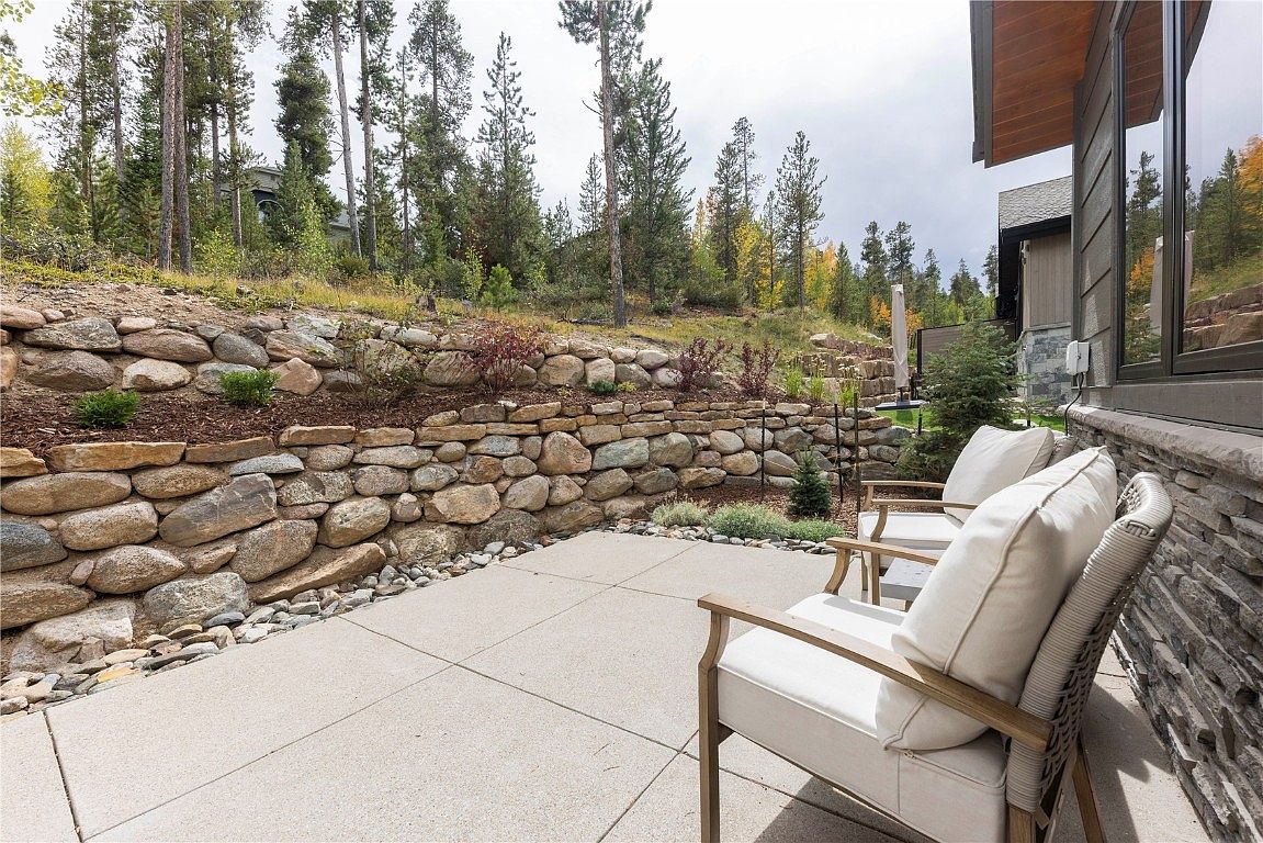 This image showcases a serene patio area adjacent to a modern home. The patio features a concrete surface, a natural stone retaining wall, and comfortable outdoor seating with white cushions. The setting is surrounded by trees, creating a private and tranquil outdoor living space.