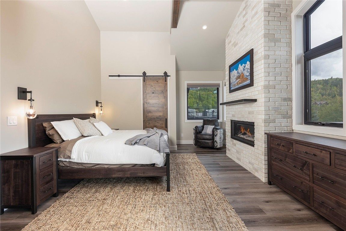 This is a primary bedroom featuring a dark wood bed frame with white linens and decorative pillows, complemented by matching nightstands. A sliding barn door adds a rustic touch, while a brick fireplace with a mounted television creates a cozy focal point. The room is well-lit with a combination of natural light from the window and modern wall sconces, creating a warm and inviting atmosphere.