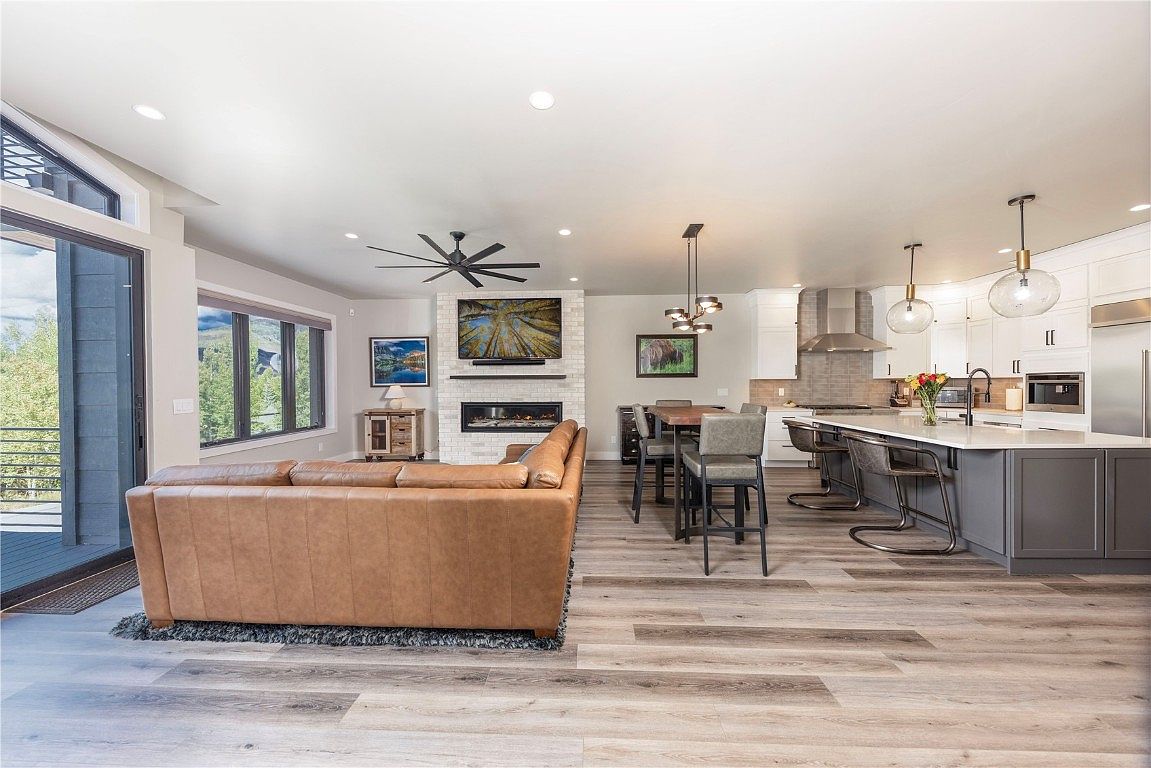 This is an interior shot of an open-concept living space, seamlessly blending the living room, dining area, and kitchen. The living room features a large leather sofa facing a fireplace with a mounted television above. The kitchen boasts a large island with bar seating and modern appliances, while the dining area includes a wooden table with upholstered chairs. The overall design is contemporary and spacious, with light wood flooring and neutral tones creating a warm and inviting atmosphere.