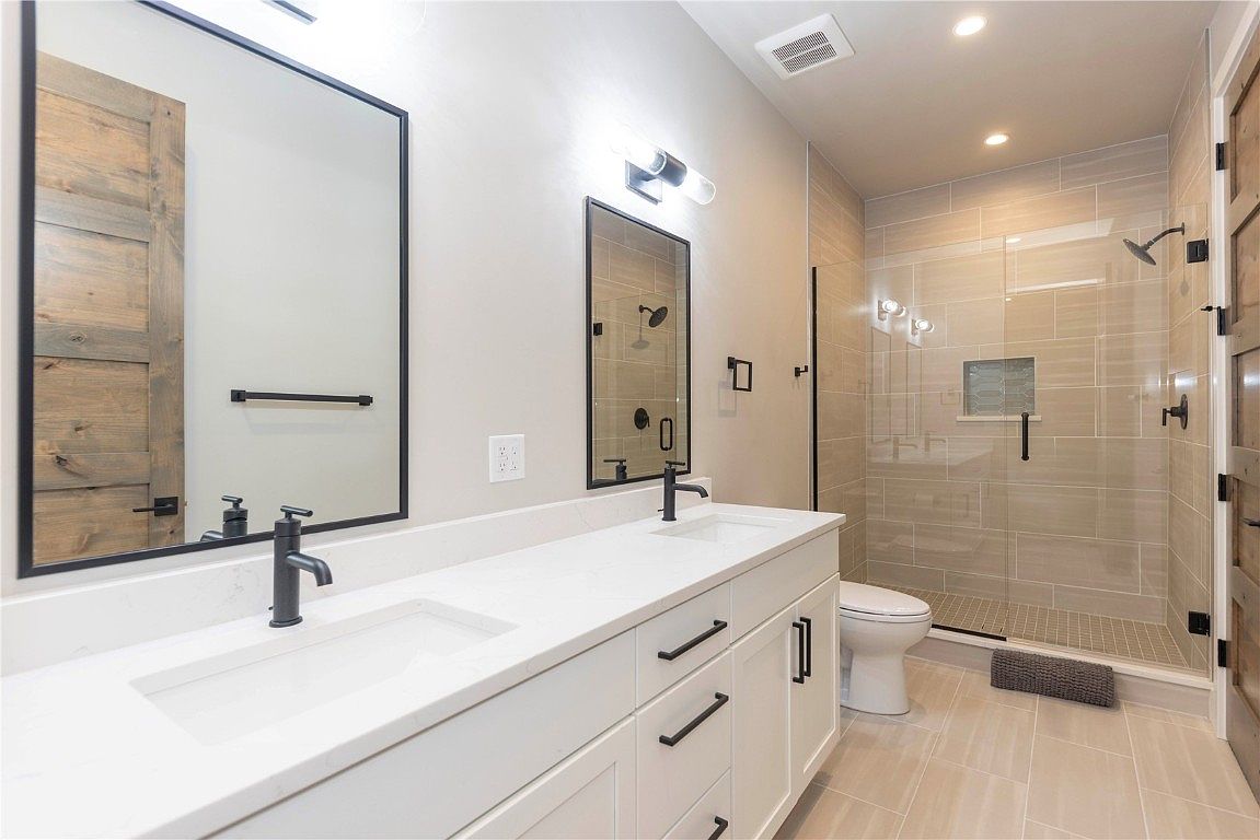 This is a well-lit primary bathroom featuring a double vanity with white cabinets and a white countertop. The bathroom has modern black fixtures, including faucets and drawer pulls, and two mirrors with black frames. A glass-enclosed shower with beige tiling is visible, and the overall design is clean and contemporary.
