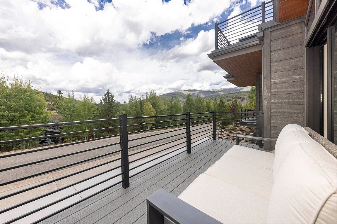 This image showcases a modern patio or deck area with sleek, horizontal railing and a comfortable-looking outdoor sofa. The deck is constructed with gray composite decking, and the view extends to a scenic landscape of trees and mountains under a partly cloudy sky. The overall impression is one of relaxation and outdoor living in a stylish setting.