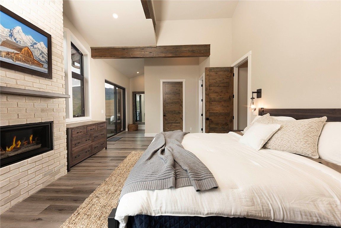 This is a primary bedroom featuring a king-size bed with white linens and decorative pillows, a brick fireplace with a mounted television, and a wooden dresser. The room has hardwood floors and wooden beams on the ceiling, adding a rustic touch. The overall impression is cozy and inviting.