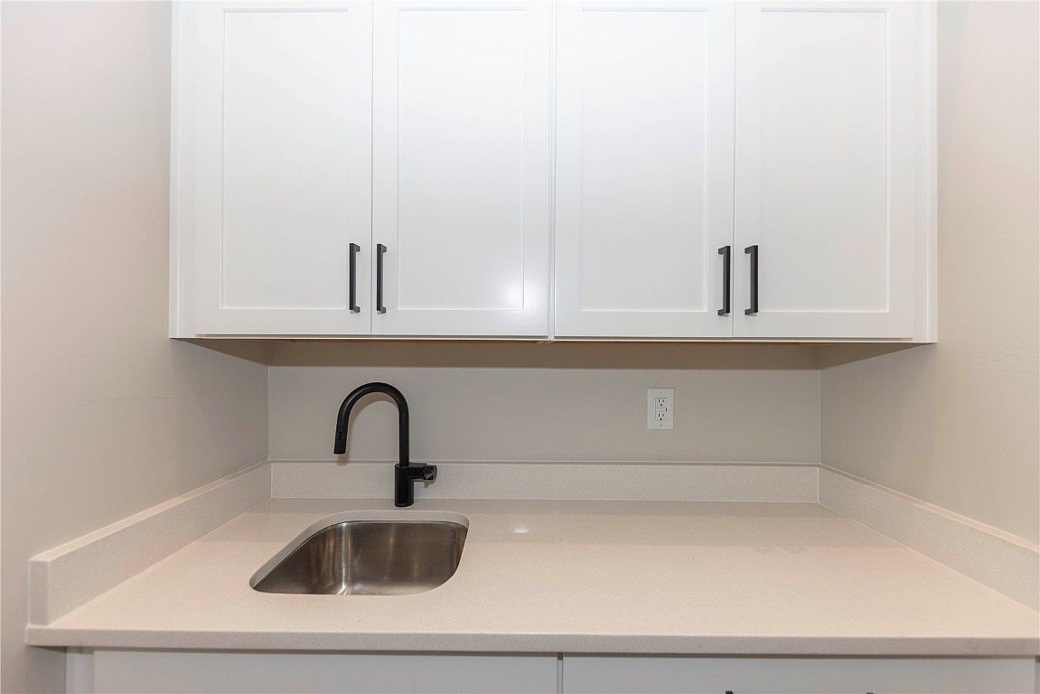 The image showcases a laundry room featuring white cabinets with dark hardware above a light-colored countertop and a stainless steel sink with a black faucet. An electrical outlet is visible on the wall, and the overall impression is clean and functional. The shot is taken from a medium perspective, focusing on the sink and cabinetry.