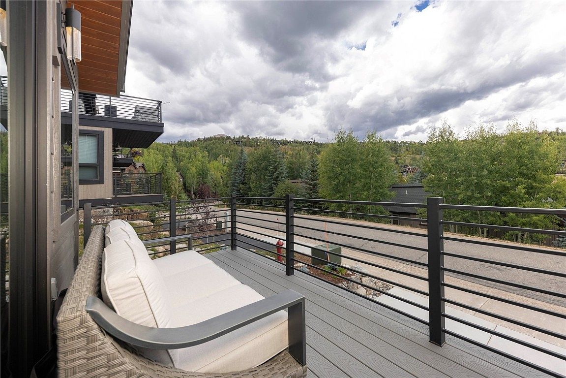 This image showcases a modern deck or balcony area, featuring a comfortable outdoor sofa with light-colored cushions. The deck is constructed with gray composite decking and surrounded by a sleek, horizontal metal railing. The view beyond the railing includes a residential street, trees, and a cloudy sky, creating a serene and inviting outdoor living space.