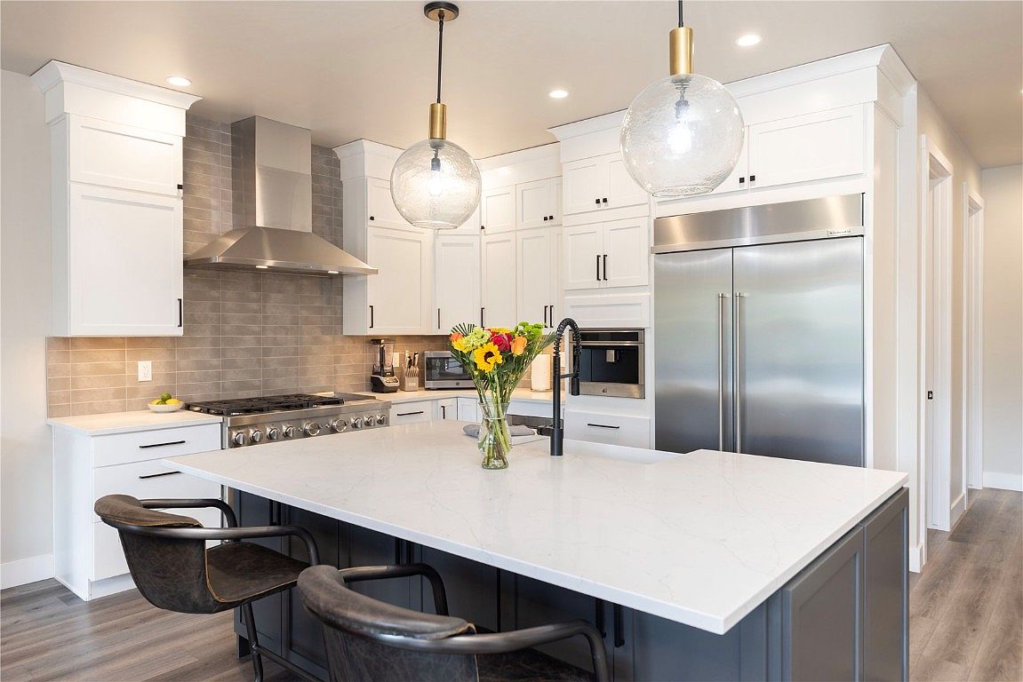 This is a bright and modern kitchen featuring white cabinetry, stainless steel appliances, and a large island with a white countertop. Two bar stools are placed at the island, and pendant lights hang above. The kitchen has a clean and contemporary style, with a focus on functionality and aesthetics.