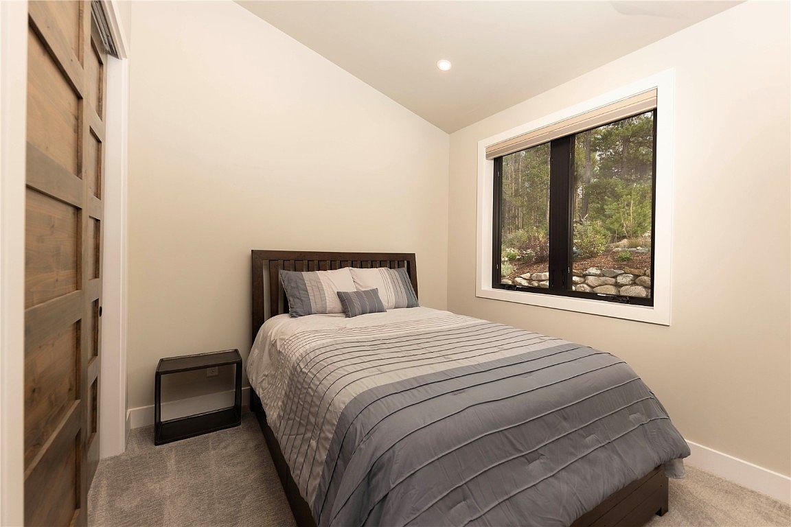This is a cozy guest bedroom featuring a wooden-framed bed with striped bedding, a dark wood nightstand, and a window overlooking a wooded area. The walls are painted in a neutral tone, and the room is carpeted, creating a warm and inviting atmosphere. A wooden sliding door adds a rustic touch to the space.