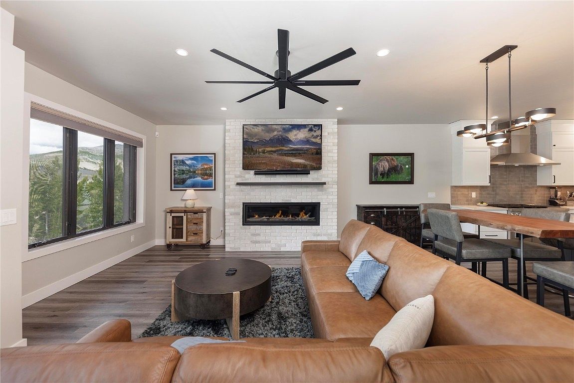 This is an interior shot of a living room featuring a large leather sectional sofa, a round coffee table on a gray rug, and a fireplace with a television mounted above it. The room has a modern aesthetic with a neutral color palette, complemented by natural light streaming through the windows and a view of the mountains. The space is designed for comfort and relaxation.
