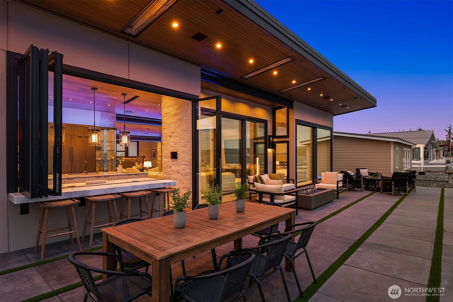 This image showcases a luxurious outdoor patio area, seamlessly blending indoor and outdoor living. A large wooden dining table with modern chairs sits prominently, while a bar area with stools connects to the interior through a folding window. The patio features sleek concrete pavers with grass inlays, and comfortable lounge seating surrounds a modern fire pit, creating an inviting space for relaxation and entertainment.