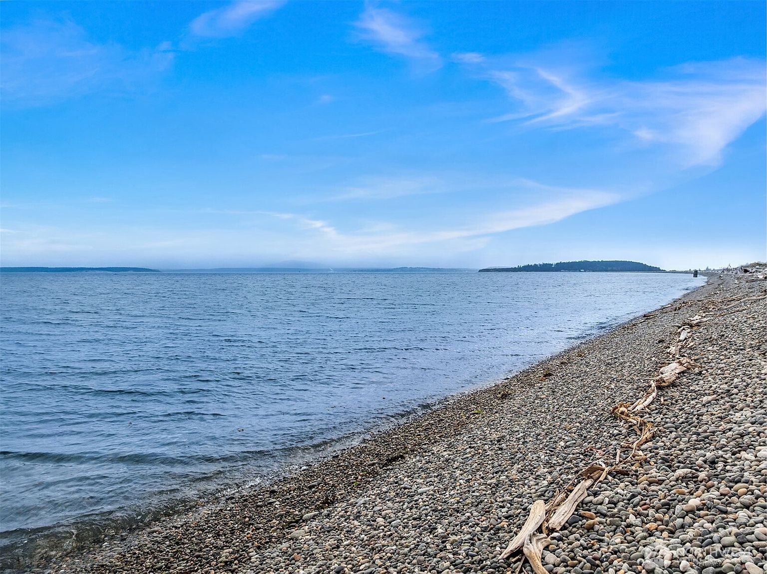The image showcases a serene beach scene with a pebble-covered shoreline meeting the calm, blue water. Driftwood is scattered along the beach, adding a natural, rustic element. In the distance, a small island is visible under a clear, blue sky with wispy clouds, creating a peaceful and inviting atmosphere.