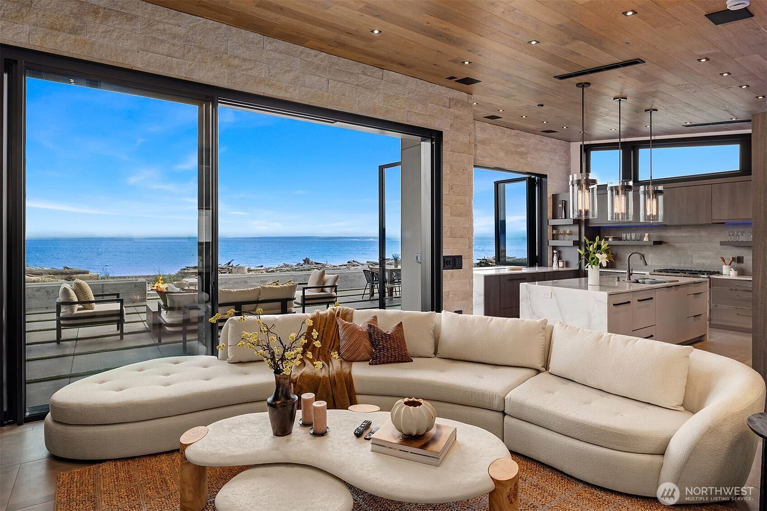 This is an interior shot of a luxurious living room with an open floor plan that seamlessly connects to the kitchen and an outdoor patio. The living room features a large, curved sectional sofa, a unique coffee table, and a textured rug. Expansive sliding glass doors offer stunning ocean views, enhancing the room's appeal and creating a serene atmosphere.