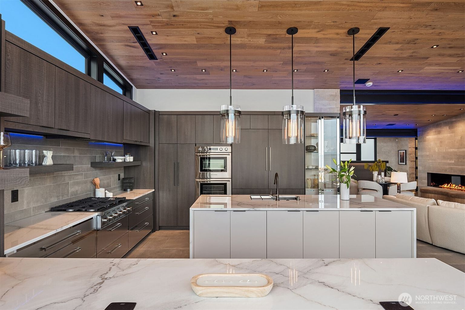 This is a modern kitchen with dark wood cabinets, stainless steel appliances, and a large island with a marble countertop. Three pendant lights hang above the island, and the ceiling is made of wood planks with recessed lighting. The kitchen opens to a living area with a fireplace, creating an open and inviting space.