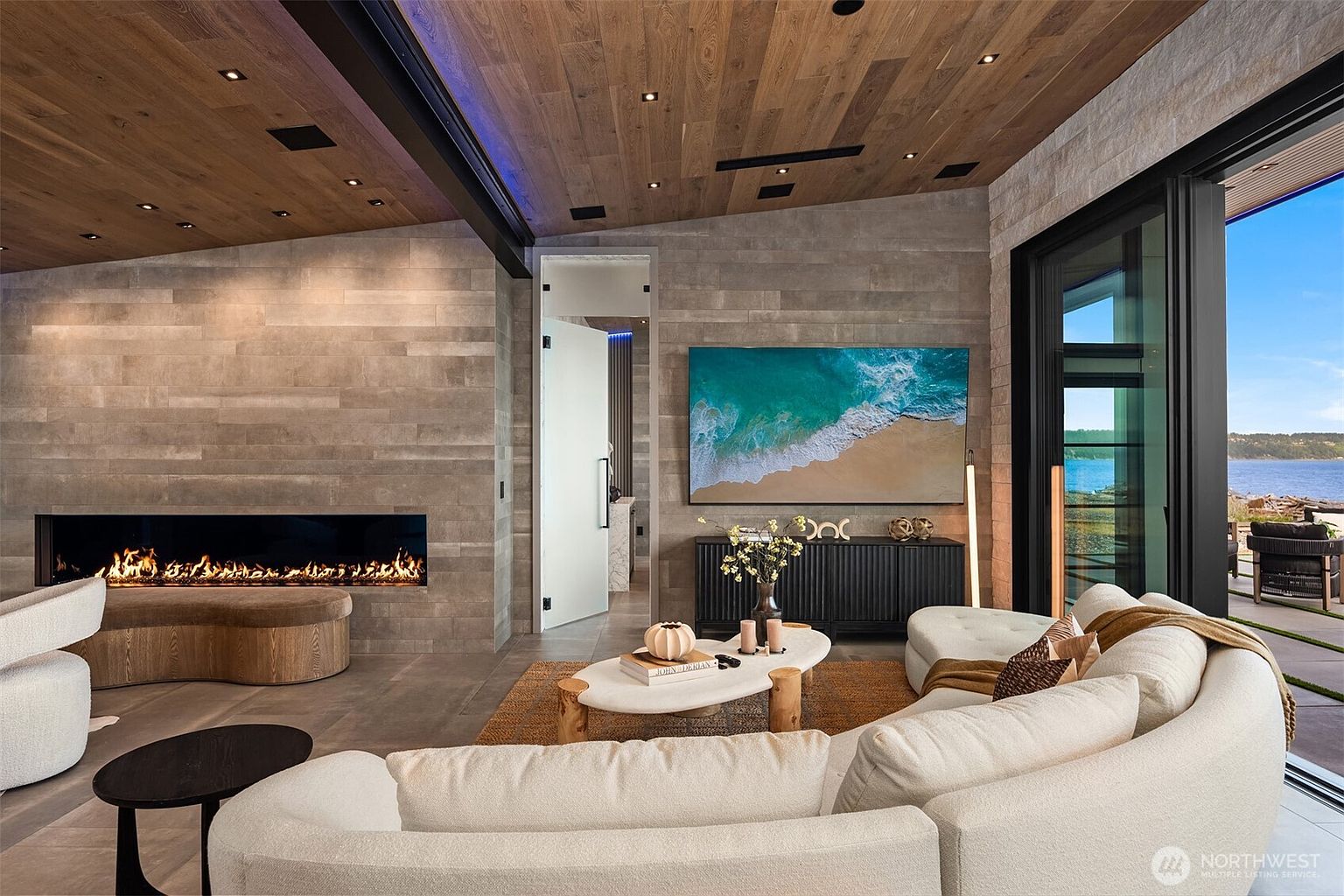 This is a luxurious living room featuring a modern fireplace, a large television mounted above a sleek console, and a comfortable curved sofa. The room has a neutral color palette with wood accents on the ceiling and walls, creating a warm and inviting atmosphere. Large sliding glass doors offer a view of the water and outdoor space, enhancing the indoor-outdoor living experience.