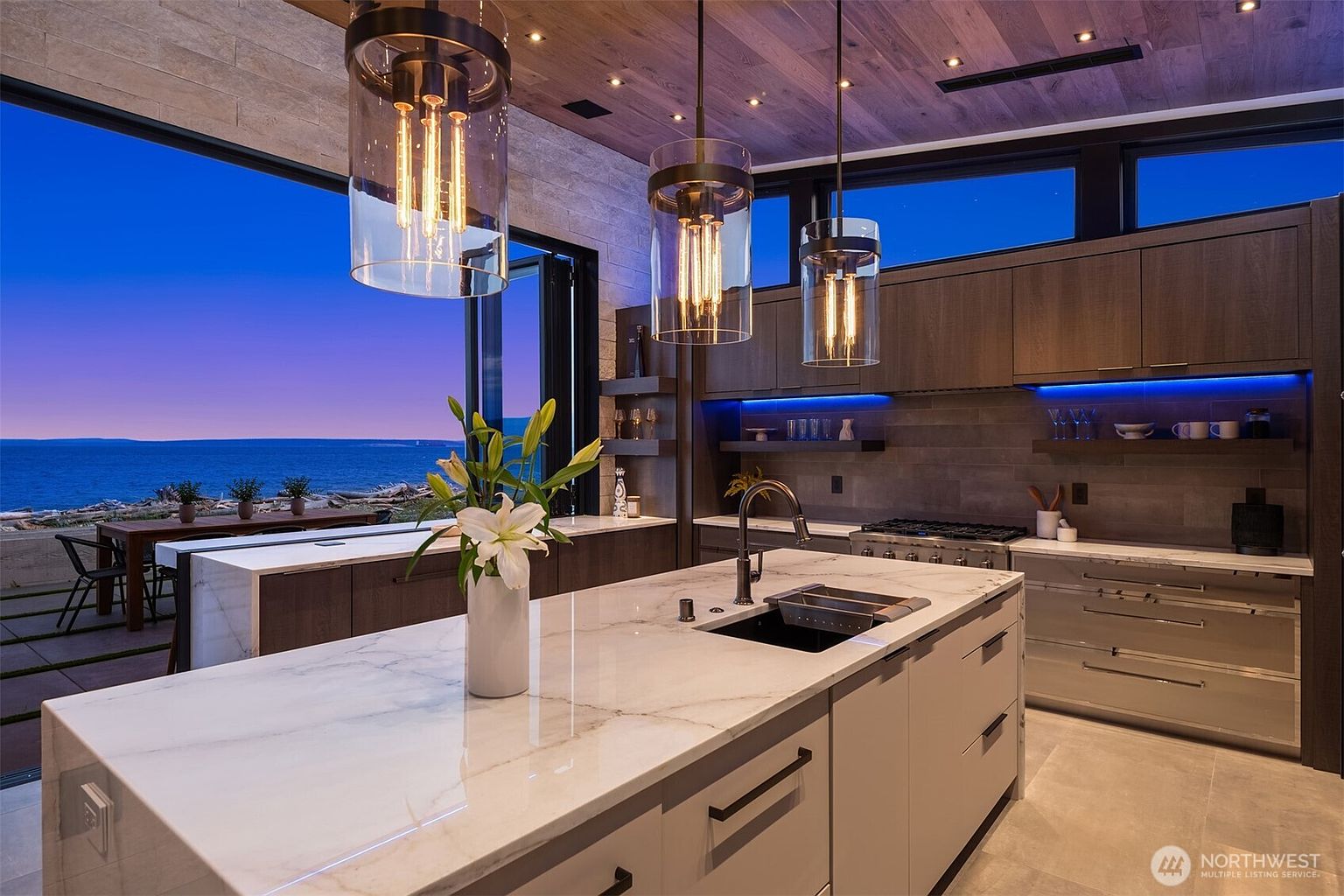 This is a modern kitchen with a large island featuring a marble countertop and a black sink. The kitchen has wooden cabinets with blue under-cabinet lighting and stainless steel appliances. Large windows offer a view of the ocean at dusk, and pendant lights hang above the island, creating a warm and inviting atmosphere.