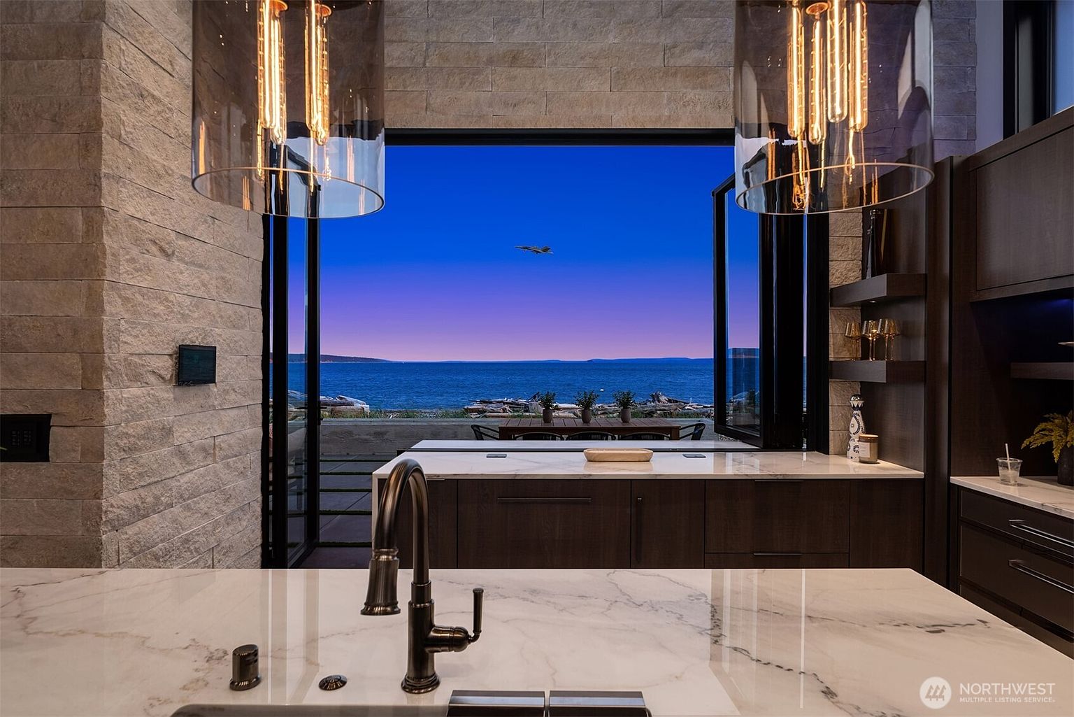This interior shot showcases a modern kitchen with a focus on the stunning view. The kitchen features a large island with a marble countertop and dark wood cabinetry. Floor-to-ceiling windows open to reveal a breathtaking ocean view with a plane flying in the distance, creating a seamless connection between the indoor space and the natural landscape.