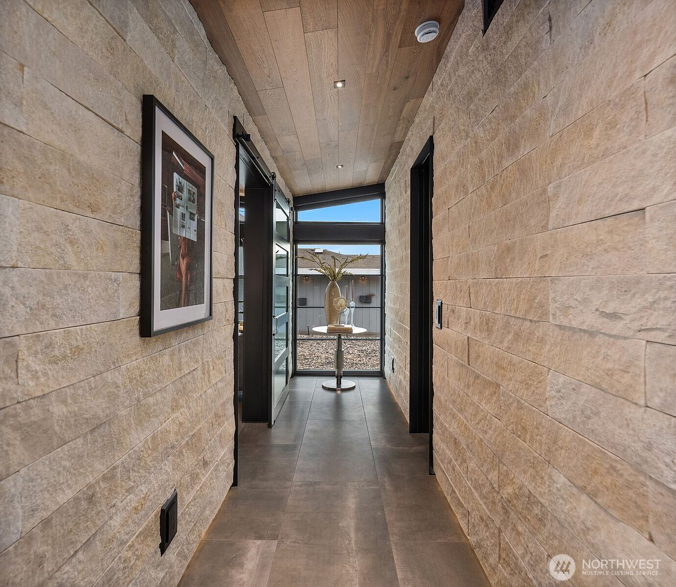 This interior shot showcases a modern hallway with stone-tiled walls and a wooden plank ceiling, creating a textured and sophisticated aesthetic. A framed artwork adorns one wall, while a glass-paneled door at the end of the hallway allows natural light to flood the space, highlighting a decorative table with a vase. The hallway's design emphasizes clean lines and a neutral color palette, contributing to a serene and upscale ambiance.