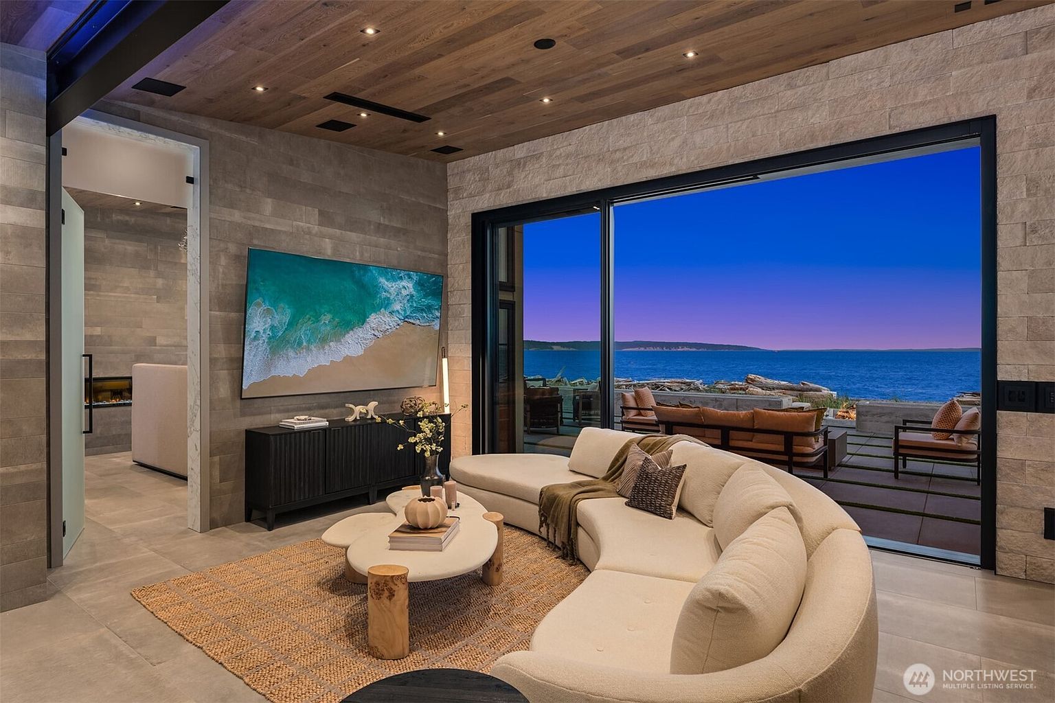 This is a stylish living room featuring a curved white sofa, a unique coffee table, and a large television displaying a beach scene. Sliding glass doors open to an outdoor patio with ocean views, creating a seamless indoor-outdoor living experience. The room is decorated with neutral tones and natural materials, giving it a modern and luxurious feel.