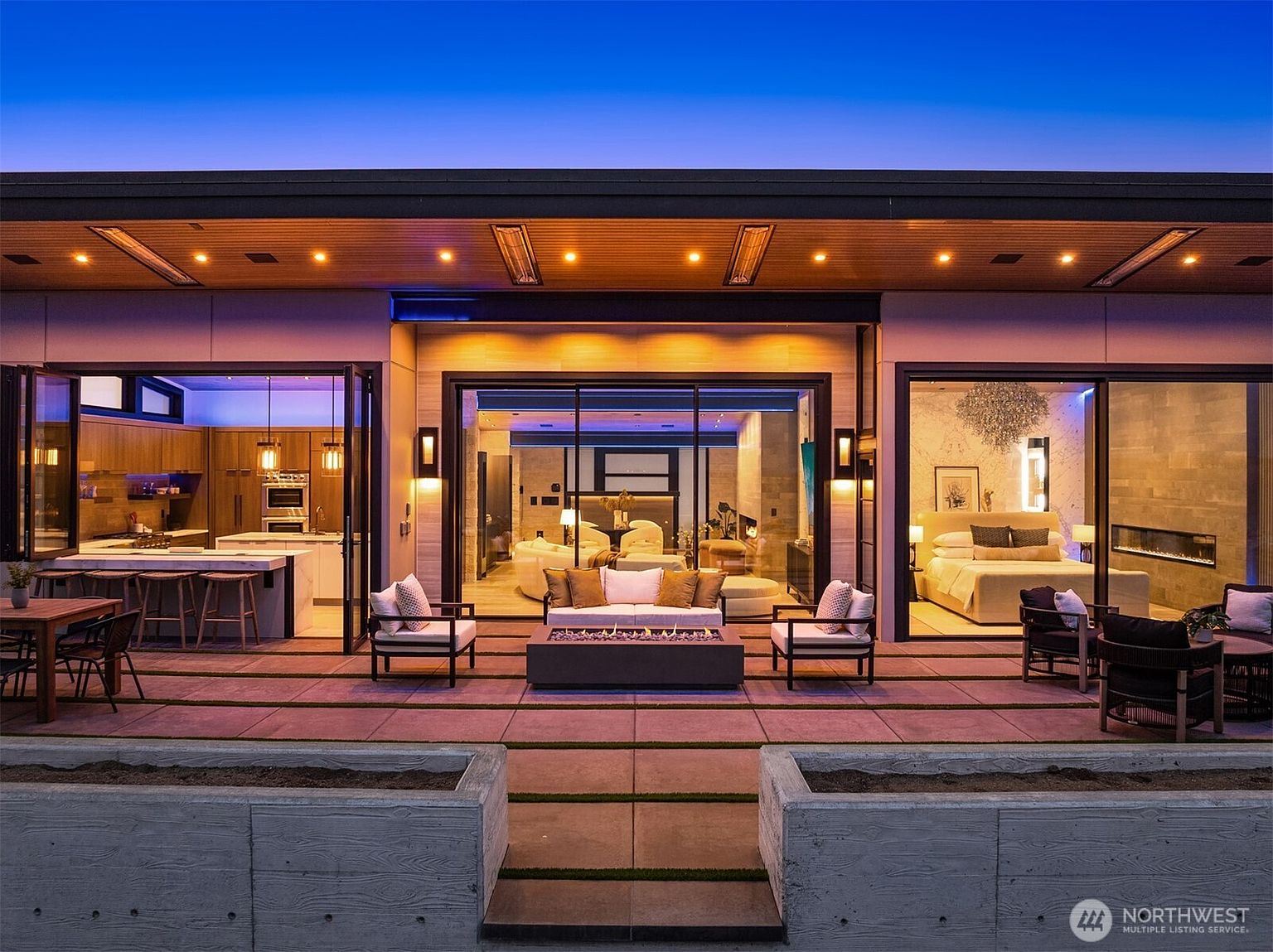 This image showcases a luxurious outdoor patio area, seamlessly blending indoor and outdoor living. The patio features comfortable seating around a modern fire pit, with large sliding glass doors opening into the home's living spaces, including a kitchen, living room, and bedroom. The design emphasizes a sophisticated and relaxed atmosphere, perfect for entertaining or enjoying quiet evenings.