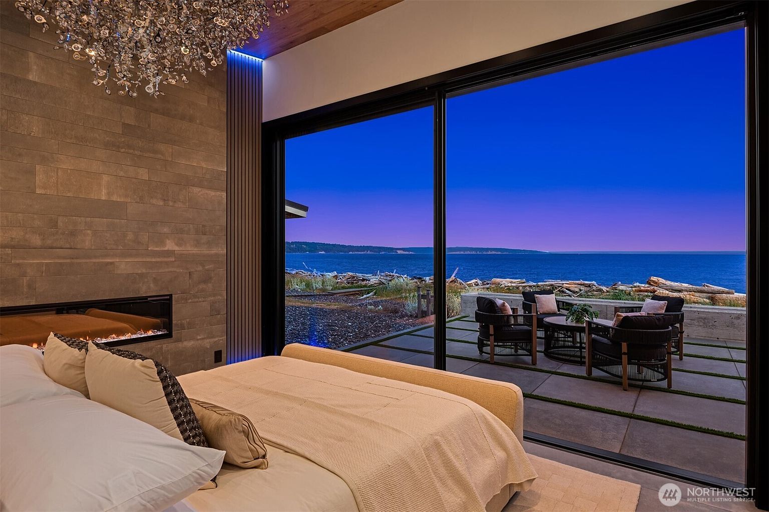 This is a luxurious primary bedroom featuring a modern fireplace and a stunning view of the ocean through large sliding glass doors. The room is elegantly furnished with a comfortable bed and a stylish chandelier, creating a serene and upscale ambiance. The outdoor patio area is visible, suggesting seamless indoor-outdoor living.