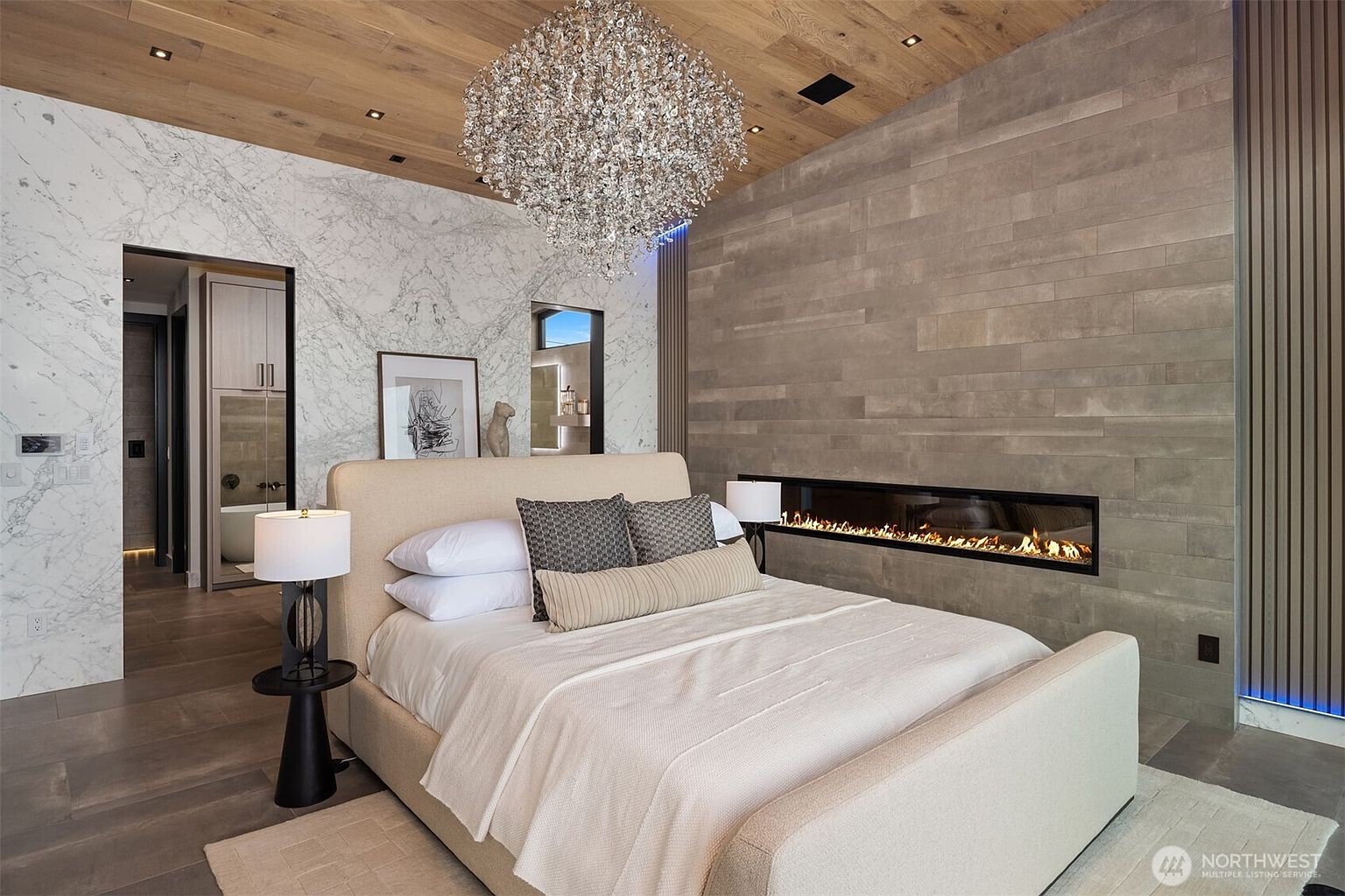 This is a luxurious primary bedroom featuring a king-size bed with neutral-toned linens and decorative pillows. The room boasts a modern fireplace set within a textured wall, a statement chandelier, and a wood-paneled ceiling. An open doorway leads to an ensuite bathroom, enhancing the sense of spaciousness and high-end design.