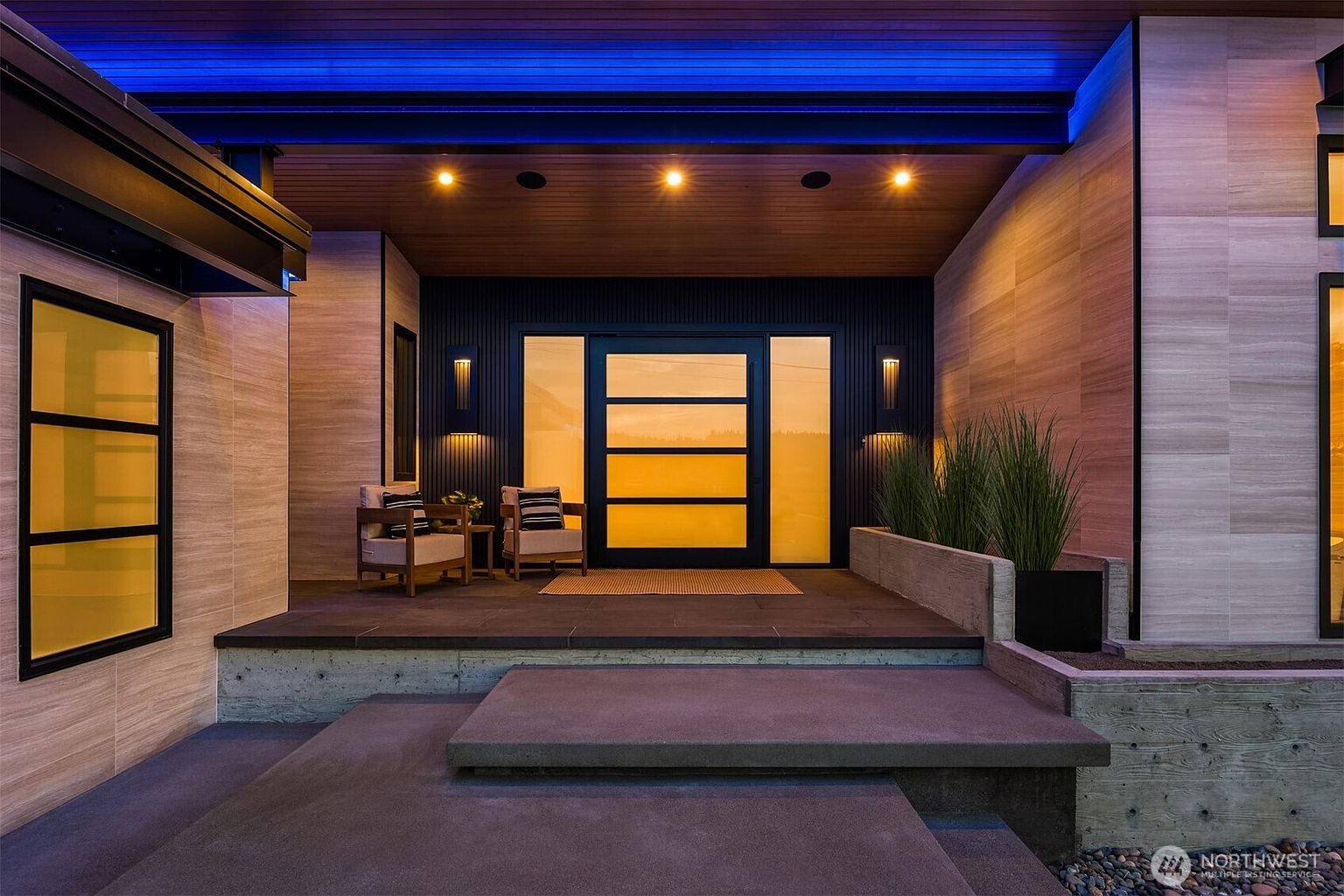 This image showcases the entryway of a modern home, featuring a sleek, dark-framed glass door flanked by vertical wall sconces. The entrance is elevated with concrete steps leading to a platform with outdoor seating, complemented by minimalist landscaping. The overall design emphasizes clean lines and a sophisticated, welcoming atmosphere.