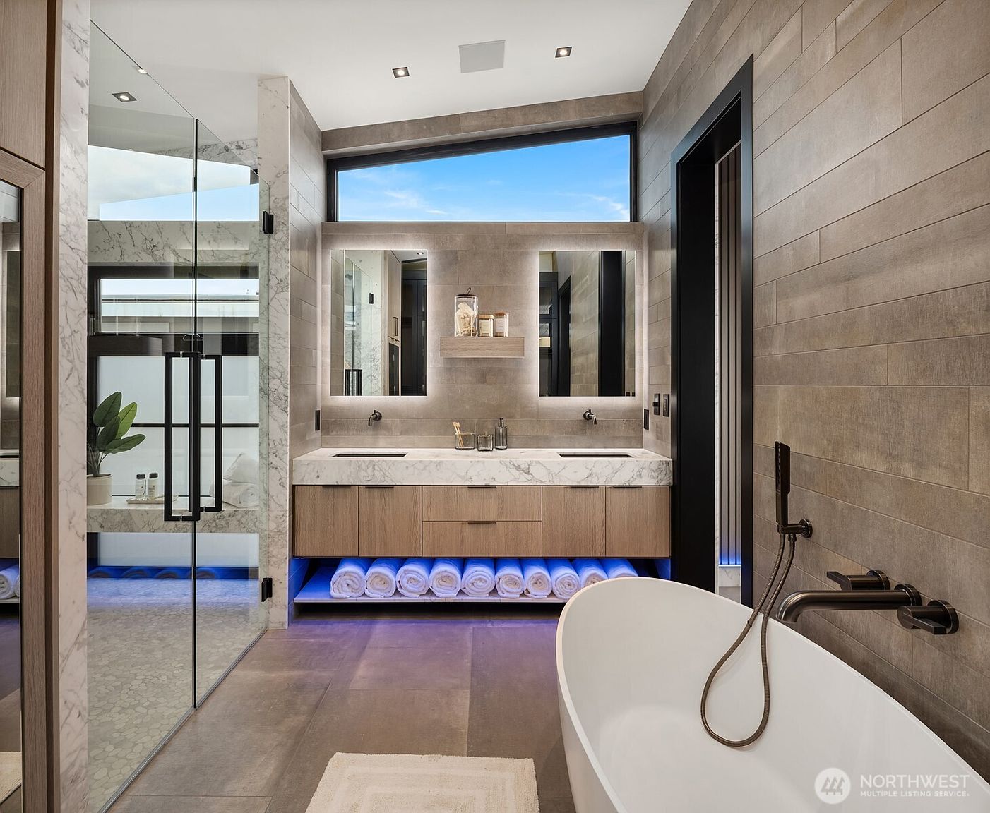 This is a luxurious primary bathroom featuring a double vanity with a marble countertop and wooden cabinets. The space is illuminated by modern, backlit mirrors and under-cabinet lighting, creating a spa-like ambiance. A freestanding bathtub with sleek fixtures adds to the room's contemporary design, while the glass-enclosed shower provides a clear view of the space.