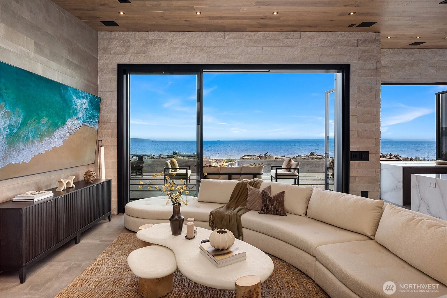 This is an interior shot of a luxurious living room with a stunning ocean view. The room features a large, curved sectional sofa, a unique coffee table with accompanying stools, and a modern entertainment center with a large screen TV. The open floor plan and natural light create a serene and inviting atmosphere, perfect for relaxation and entertaining.