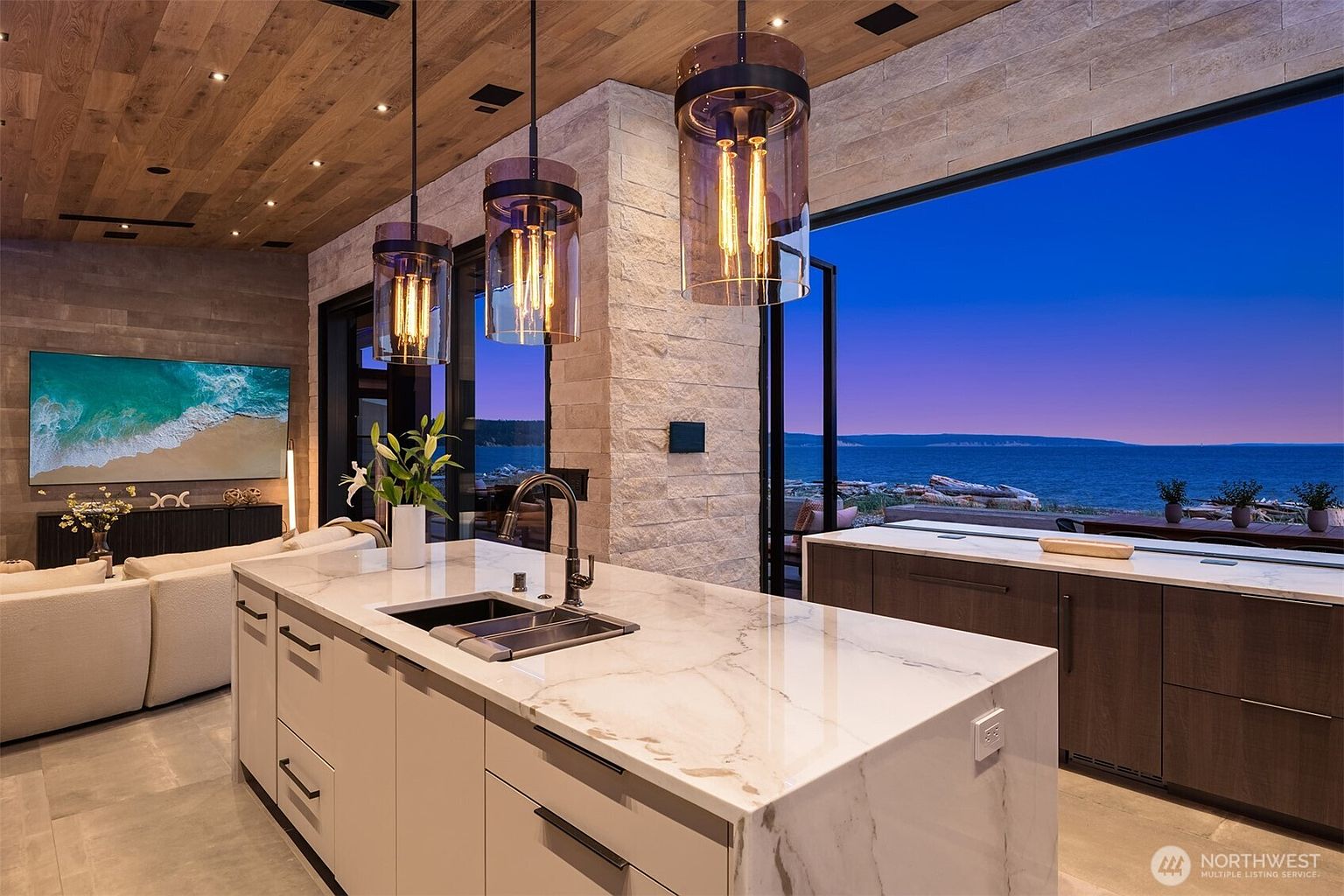 This is a beautifully designed kitchen featuring a large island with a marble countertop and a black sink. Pendant lights hang above the island, and a large window offers a stunning view of the ocean. The kitchen has a modern and luxurious feel, with a seamless blend of natural materials and contemporary design.