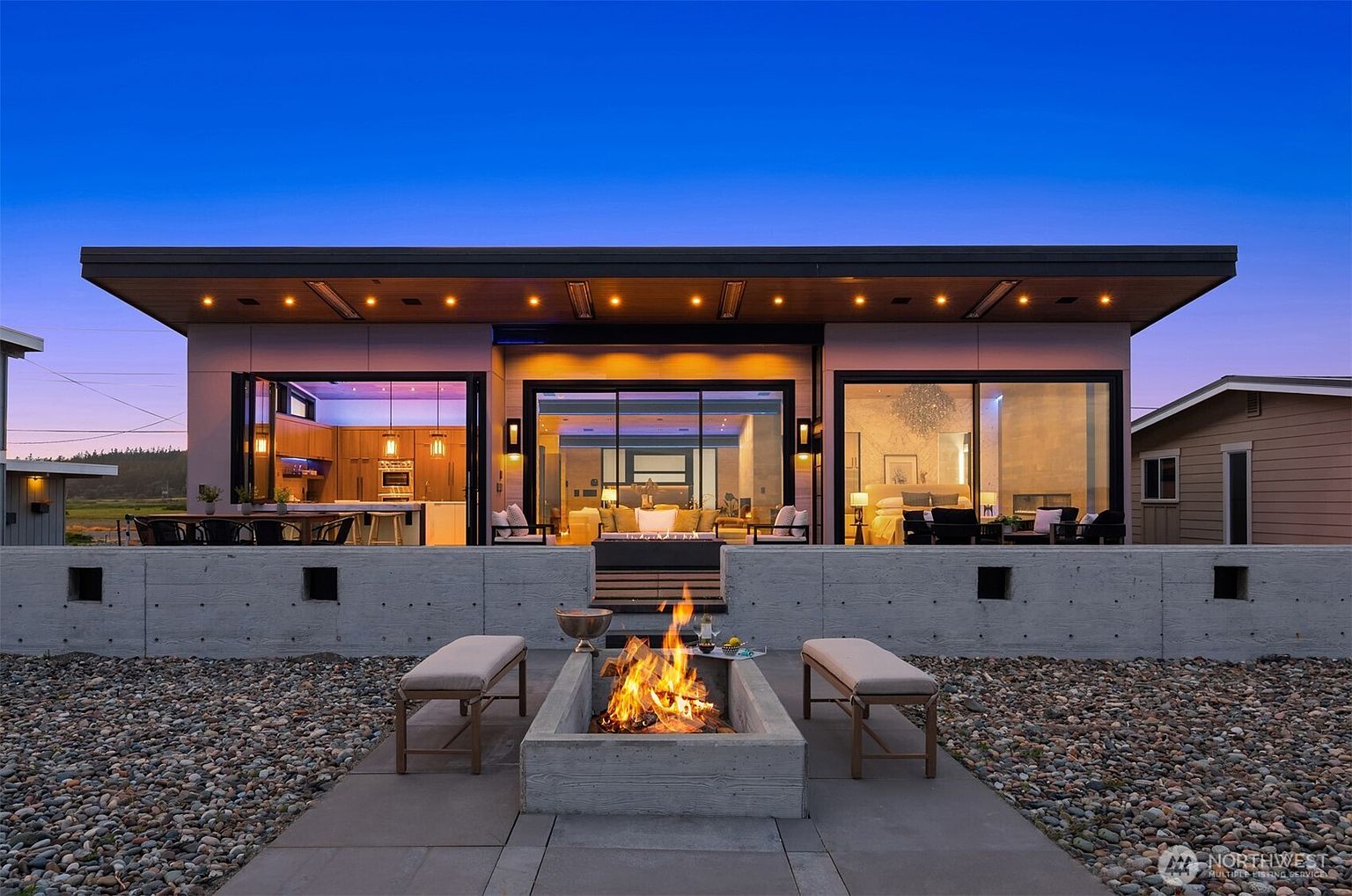 This image showcases a modern home's outdoor patio area at dusk, featuring a concrete fire pit with a lively fire as the focal point. Two benches flank the fire pit, inviting relaxation, while the home's open-concept design is visible through large sliding glass doors, revealing glimpses of the interior spaces. The patio is surrounded by decorative gravel, adding texture and visual interest to the outdoor space.