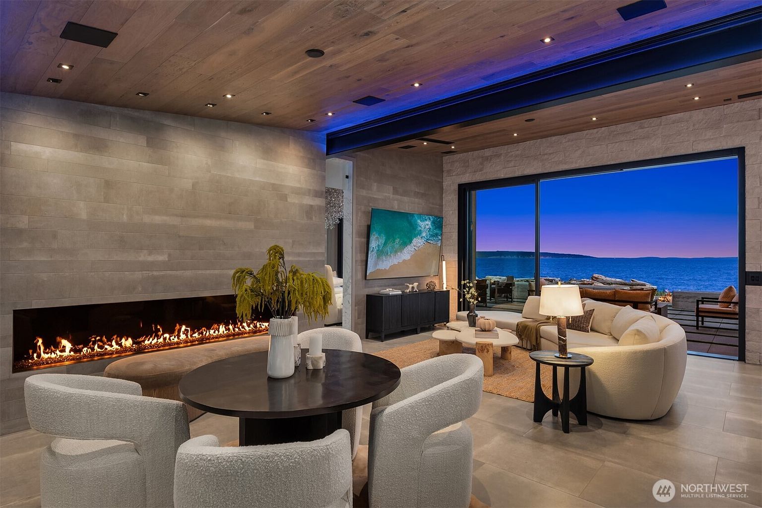 This is a luxurious living room featuring a modern fireplace, a round dining table with plush chairs, and a comfortable seating area with a view of the ocean. The room is decorated in neutral tones with wood accents, creating a warm and inviting atmosphere. The large window provides ample natural light and a stunning view.