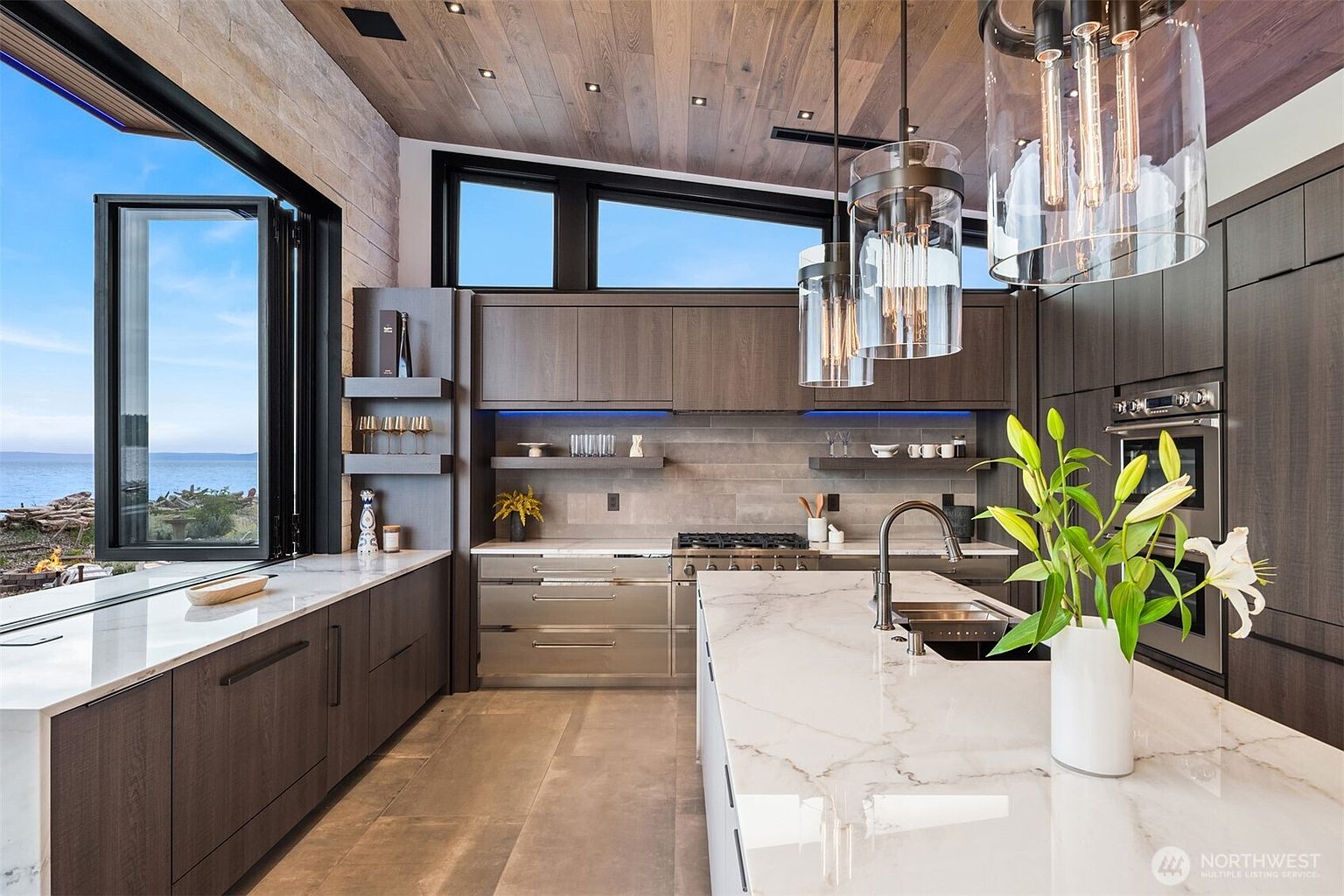This is a modern kitchen with sleek, dark wood cabinetry and a large marble island. The kitchen features stainless steel appliances, open shelving, and unique pendant lighting. A large window offers a view of the ocean, creating a luxurious and inviting space.
