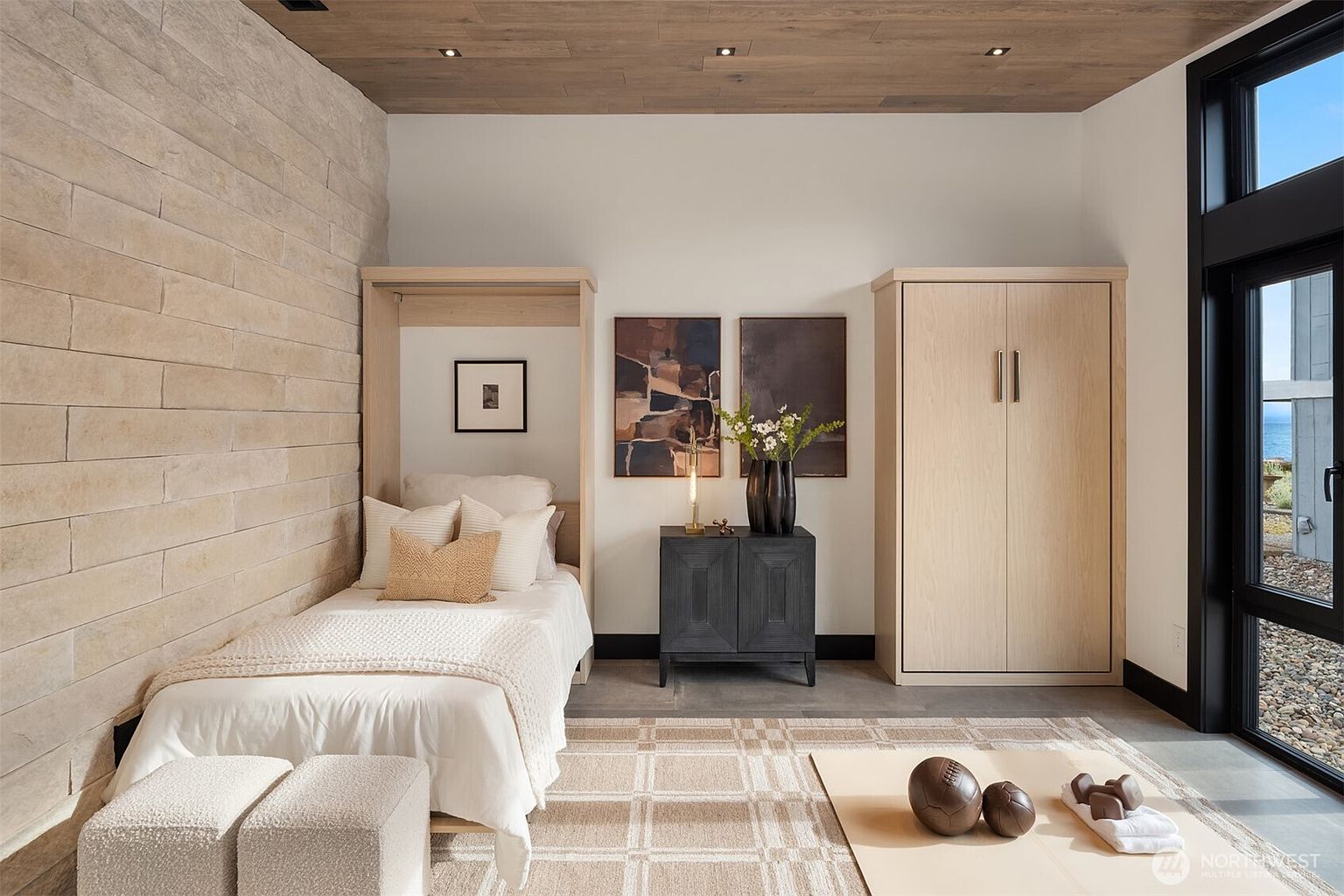 This is a well-appointed guest bedroom featuring a built-in Murphy bed with a light wood frame, complemented by a stone accent wall. The room includes a dark wood nightstand with decorative art above, a matching light wood wardrobe, and a neutral-toned rug. The space is bright and airy, with a modern aesthetic and a view to the outside.