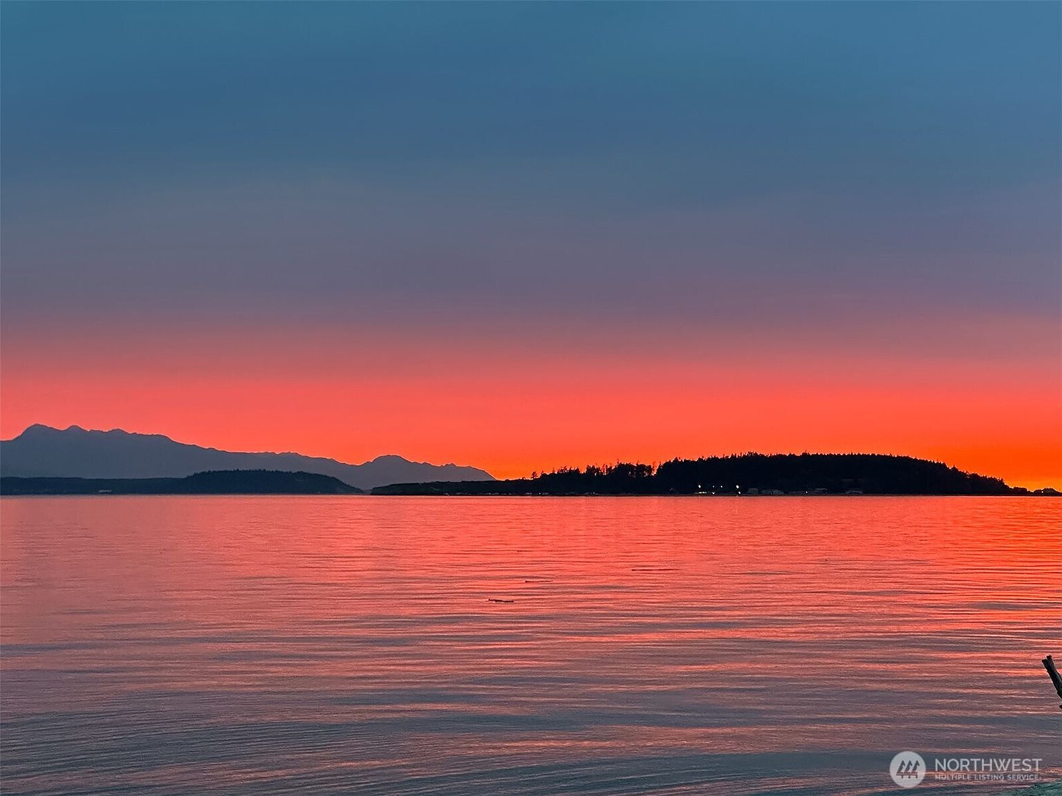 The image captures a serene sunset over a body of water, likely a sea or large lake. The sky transitions from a deep blue at the top to a vibrant orange and red near the horizon, reflecting beautifully on the water's surface. Silhouetted islands and mountains add depth to the scene, creating a peaceful and picturesque view.