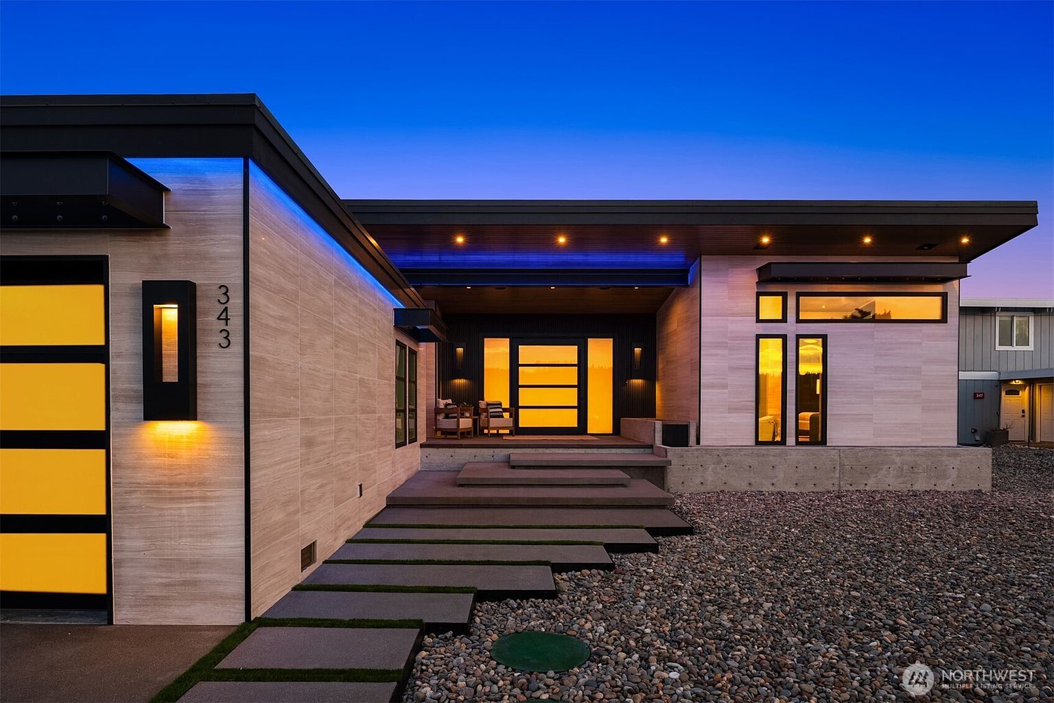 This is a front exterior view of a modern home at dusk. The house features a flat roof, a combination of light-colored stone and dark accents, and a striking front door with horizontal panels illuminated from within. The landscaping includes gravel and stepping stones leading to the entrance, creating a sleek and inviting approach.