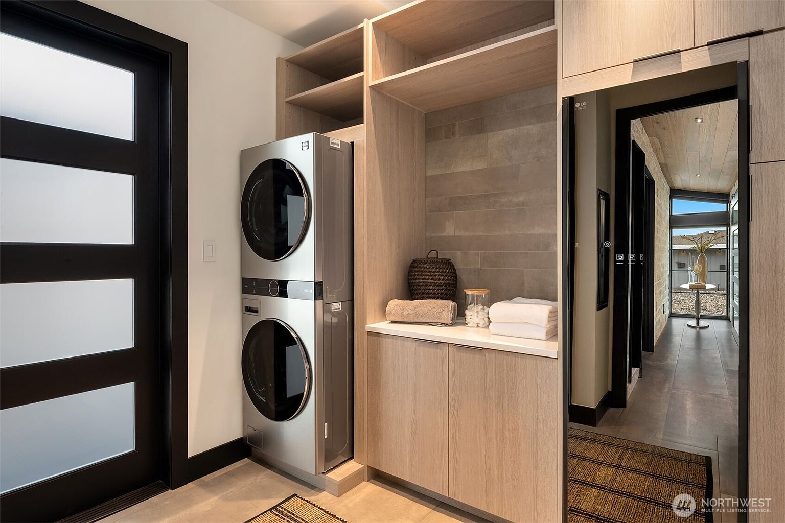 This is a well-organized laundry room featuring a stacked washer and dryer set, custom cabinetry with open shelving, and a countertop for folding. The room has a modern aesthetic with clean lines and neutral tones, creating a functional and stylish space. A doorway leads to another room, suggesting a seamless flow within the home.