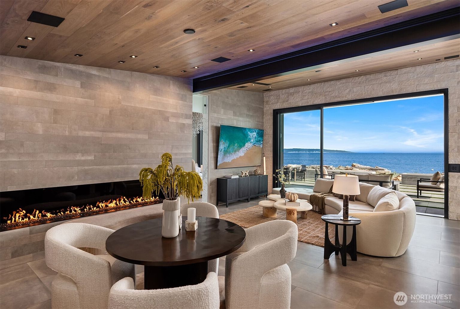 This is an interior shot of a luxurious living room featuring a modern fireplace, a round dining table with four chairs, and a comfortable seating area with a curved sofa. A large sliding glass door offers stunning ocean views, enhancing the room's appeal. The room is decorated in neutral tones with wood and stone accents, creating a sophisticated and inviting atmosphere.