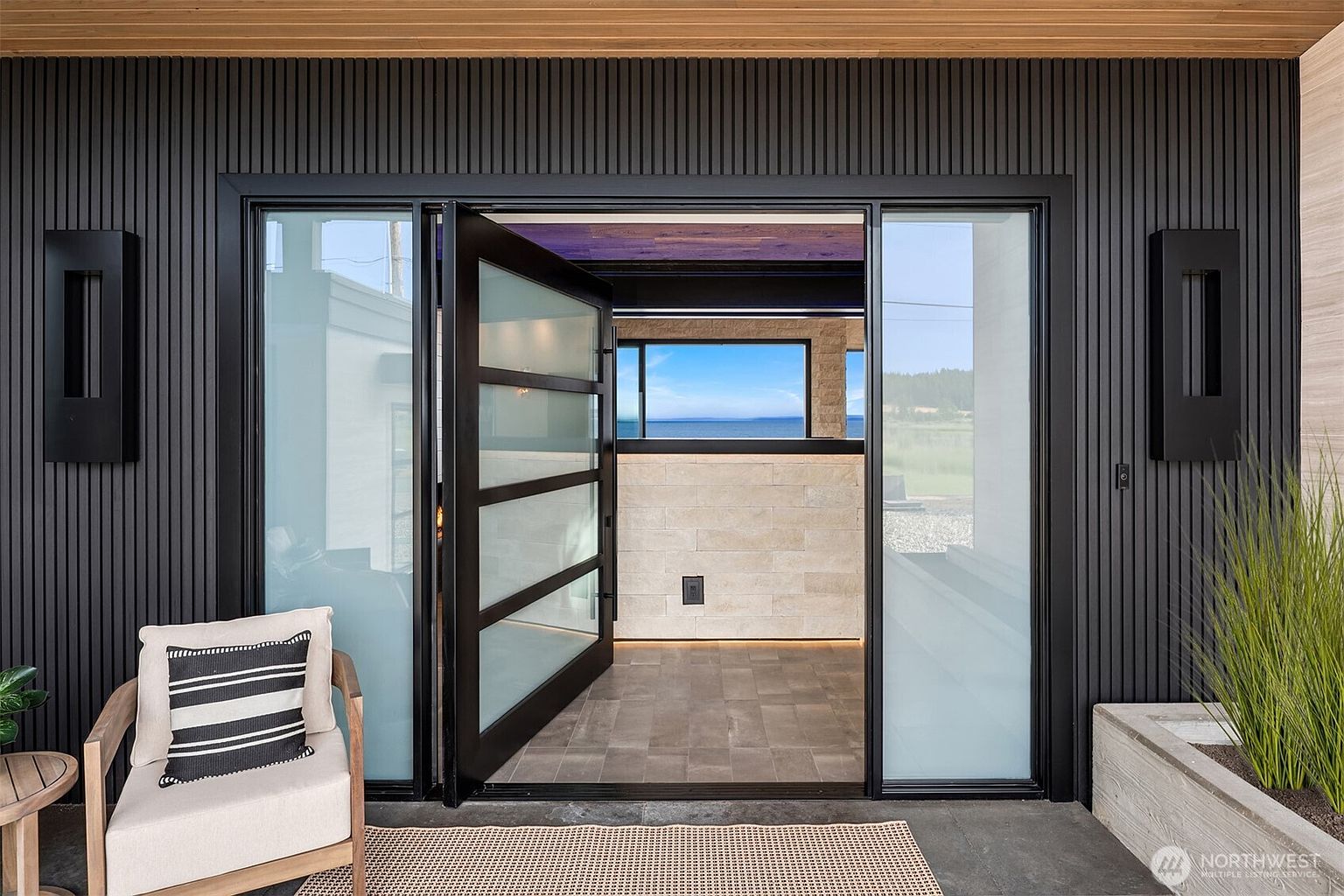 This image showcases a modern home's entryway, featuring a striking black exterior with vertical paneling and a contemporary glass door. The open door reveals a glimpse of the interior, including a view of the ocean through a window, creating an inviting and luxurious first impression. A stylish chair and planter add to the sophisticated aesthetic.