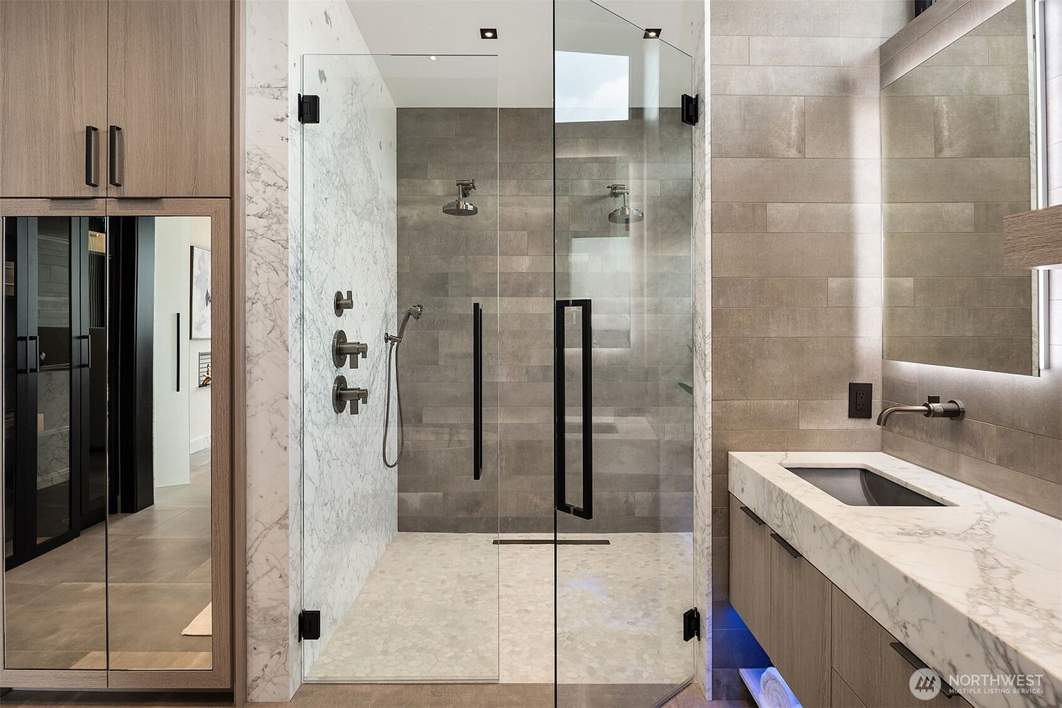 This is a modern primary bathroom featuring a glass-enclosed shower with dual shower heads and marble accents. The vanity has a marble countertop and wood-grain cabinets with under-cabinet lighting. The overall design is sleek and luxurious, creating a spa-like atmosphere.