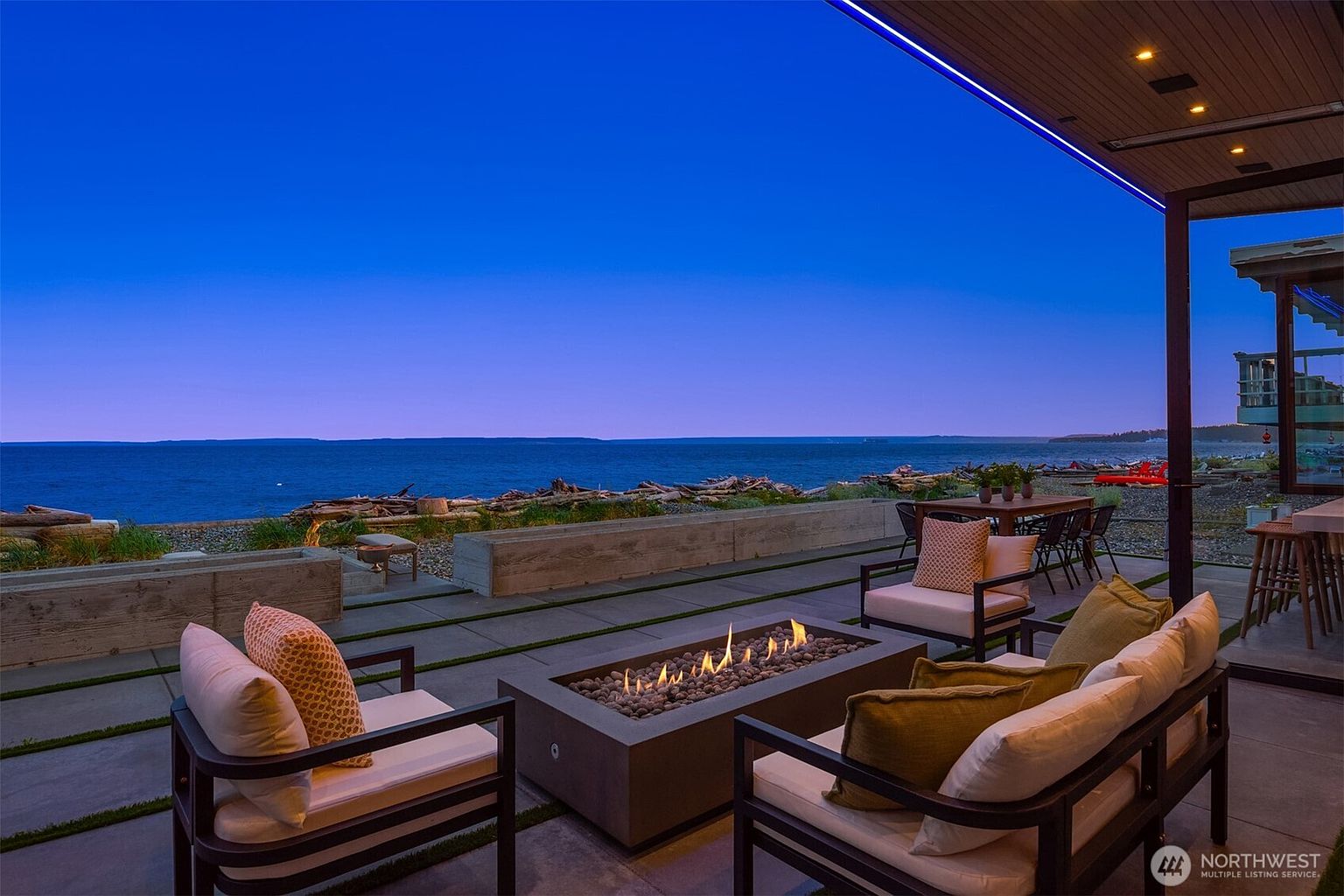 This image showcases a luxurious outdoor patio area with a modern fire pit as the focal point, surrounded by comfortable seating. The patio overlooks a scenic ocean view with driftwood scattered along the beach, creating a serene and upscale ambiance. The architectural design of the house is contemporary, with clean lines and subtle lighting enhancing the outdoor living space.