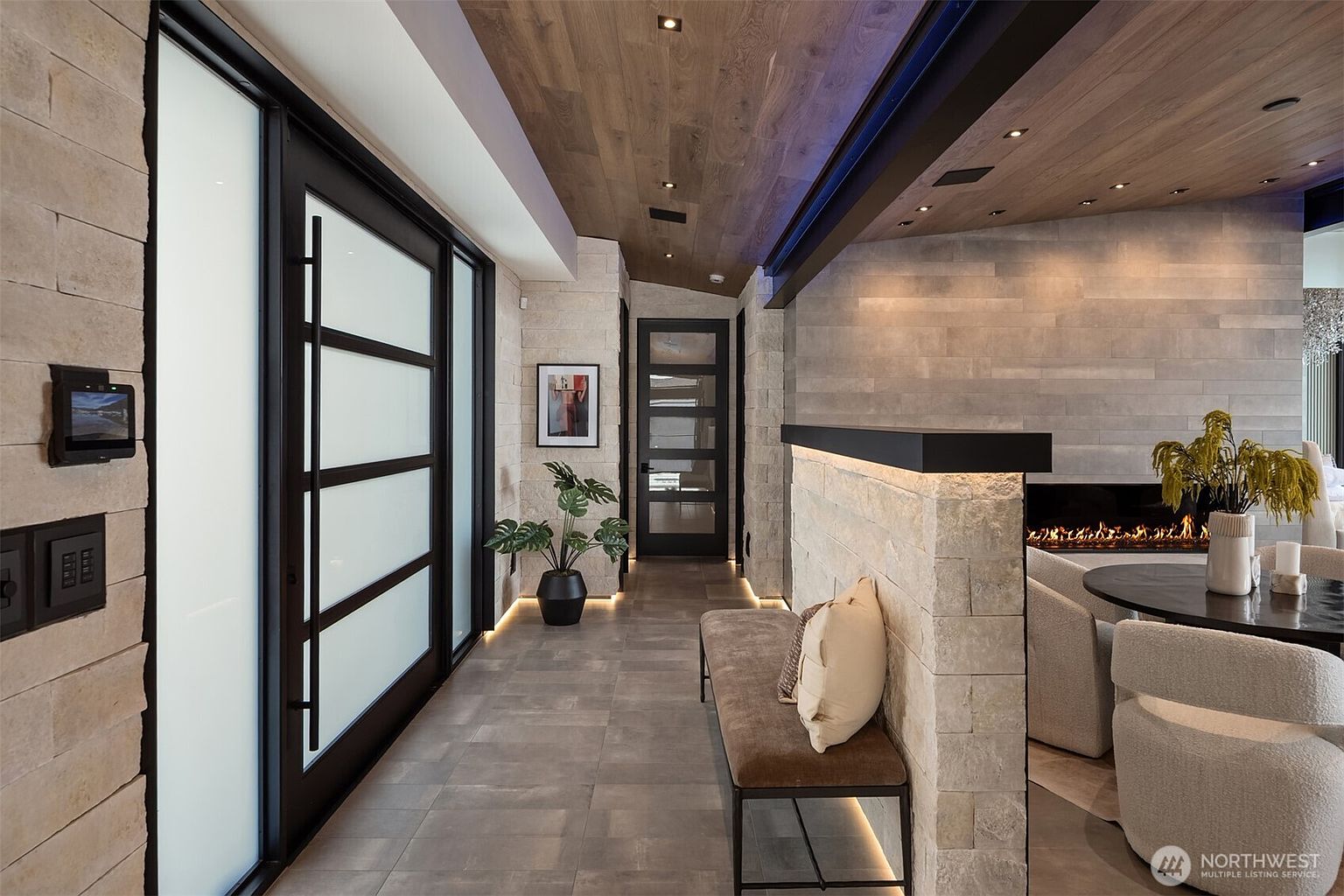 This interior shot showcases a modern hallway with a blend of textures and materials. The walls are clad in light-colored stone, complemented by a wood-paneled ceiling with recessed lighting. A sleek bench with a pillow sits against the wall, and a glimpse of a fireplace and living area can be seen in the background, creating an inviting and stylish transition space.