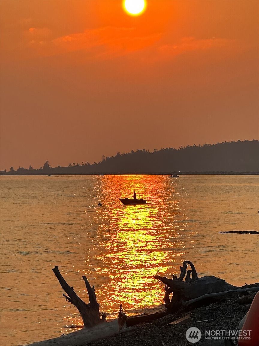 The image captures a serene sunset over a body of water, with a silhouette of a person in a small boat in the middle ground. The sun's reflection creates a golden path on the water's surface, leading the eye towards the horizon. In the foreground, driftwood adds texture and depth to the scene, enhancing the natural beauty and tranquility of the location.