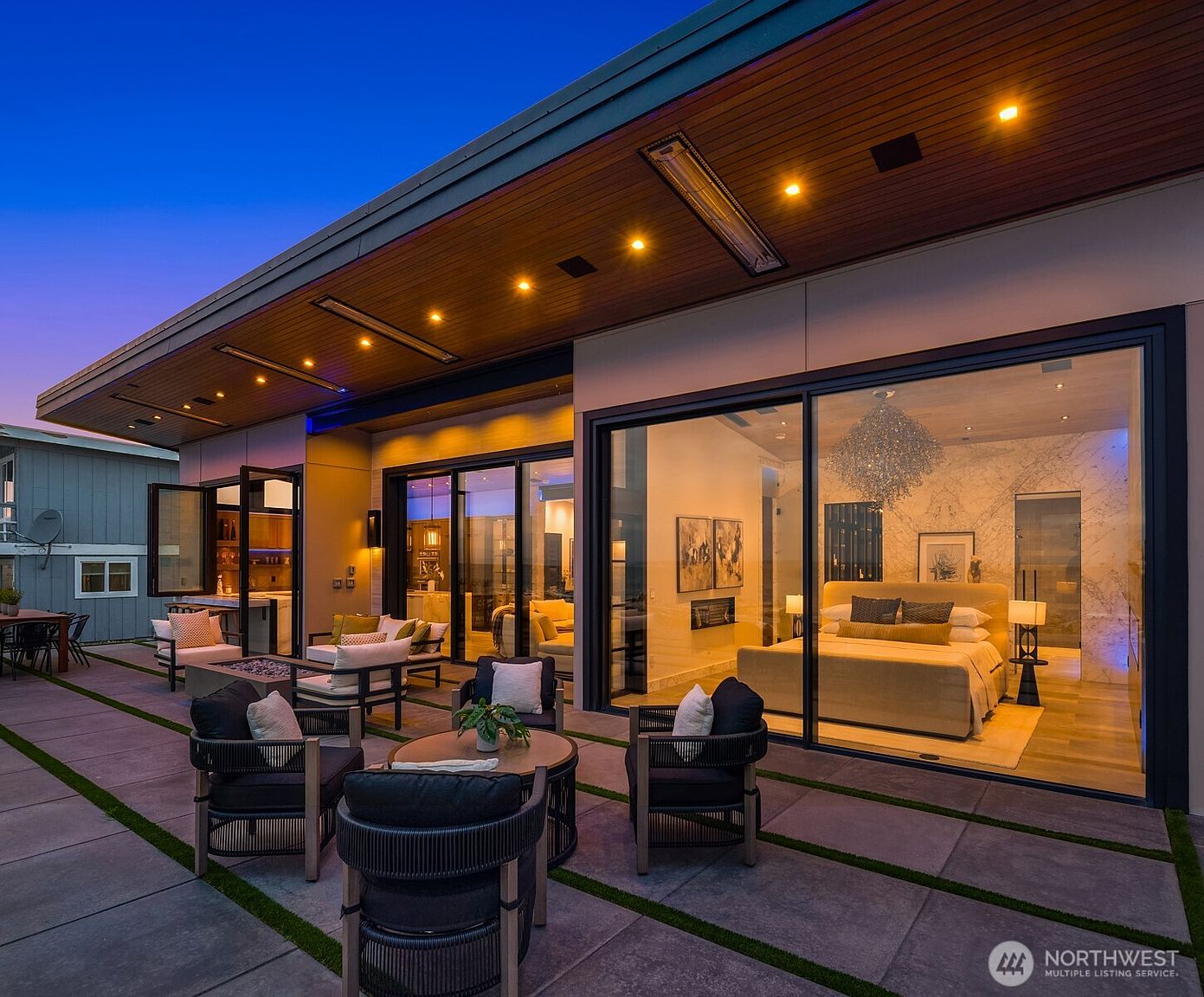 This image showcases a luxurious outdoor patio area with modern design elements. The patio features stylish outdoor furniture, including chairs and tables, arranged on a paved surface with grass accents. Large sliding glass doors provide seamless access to the interior, revealing a glimpse of a beautifully decorated bedroom with a chandelier.