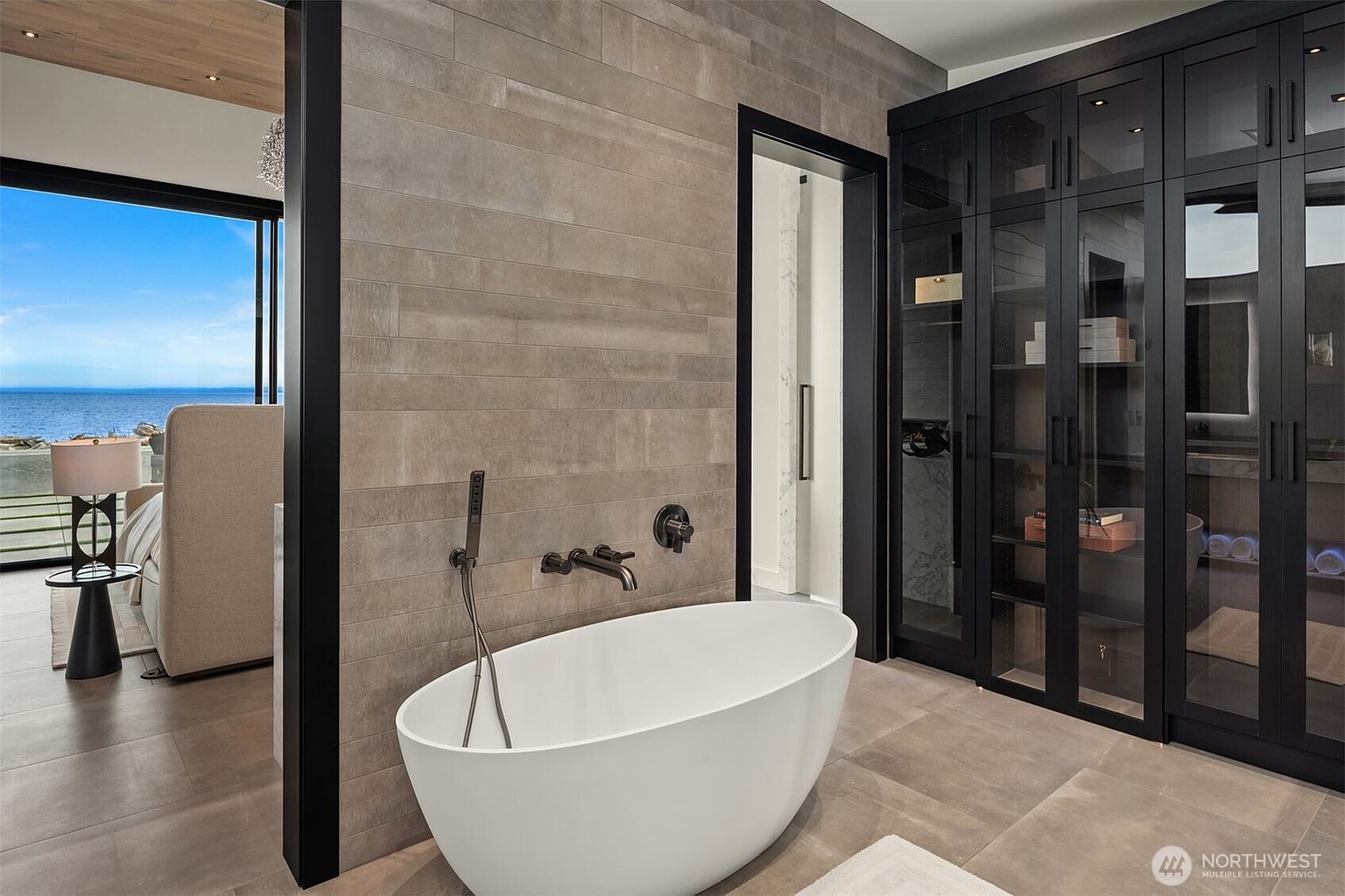 This is a luxurious primary bathroom featuring a freestanding white bathtub with modern fixtures. The walls are covered in horizontal stone-look tiles, and a black-framed glass door leads to a shower area. A large black cabinet with glass doors provides ample storage, and the flooring is a matching stone tile. The perspective is from the doorway, showcasing the tub and the view to the bedroom.