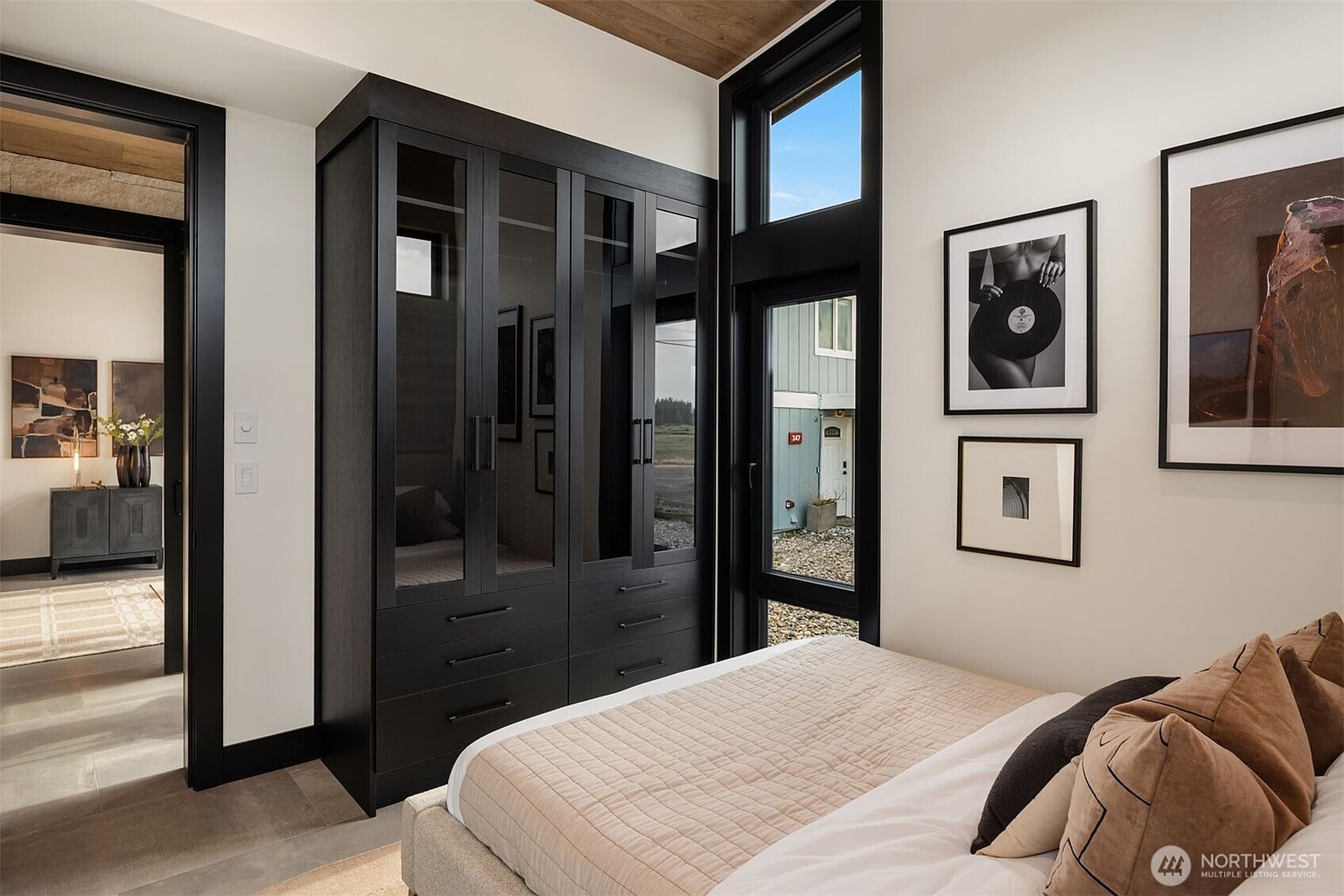 This is a bedroom featuring a dark wood wardrobe with glass doors and drawers, a bed with neutral-toned bedding and pillows, and framed artwork on the wall. A black-framed window provides natural light and a view to the outside. The room has a modern and sophisticated aesthetic.