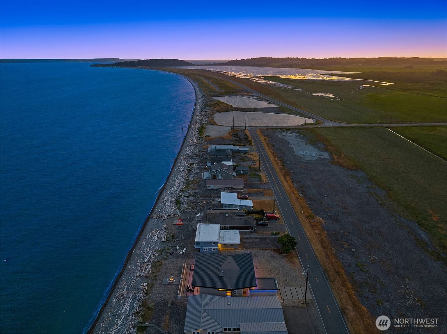 This aerial view showcases a row of beachfront houses nestled along a pebble beach, with the vast ocean on one side and a road and marshland on the other. The houses vary in style, some featuring modern designs with dark roofs and outdoor fire pits, while others have a more traditional coastal aesthetic. The image captures the unique setting and the proximity of the homes to both the water and the natural landscape.