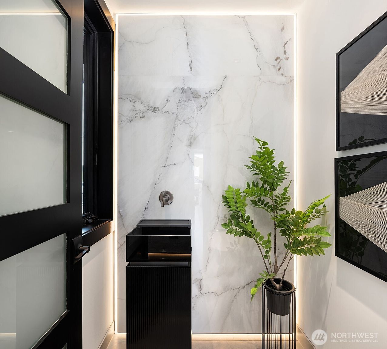 This is a modern bathroom featuring a sleek, black vanity with a wall-mounted faucet against a backdrop of large-format marble-look tiles illuminated by vertical LED strips. A potted plant adds a touch of nature, while framed artwork enhances the contemporary aesthetic. The overall impression is one of minimalist elegance and sophisticated design.