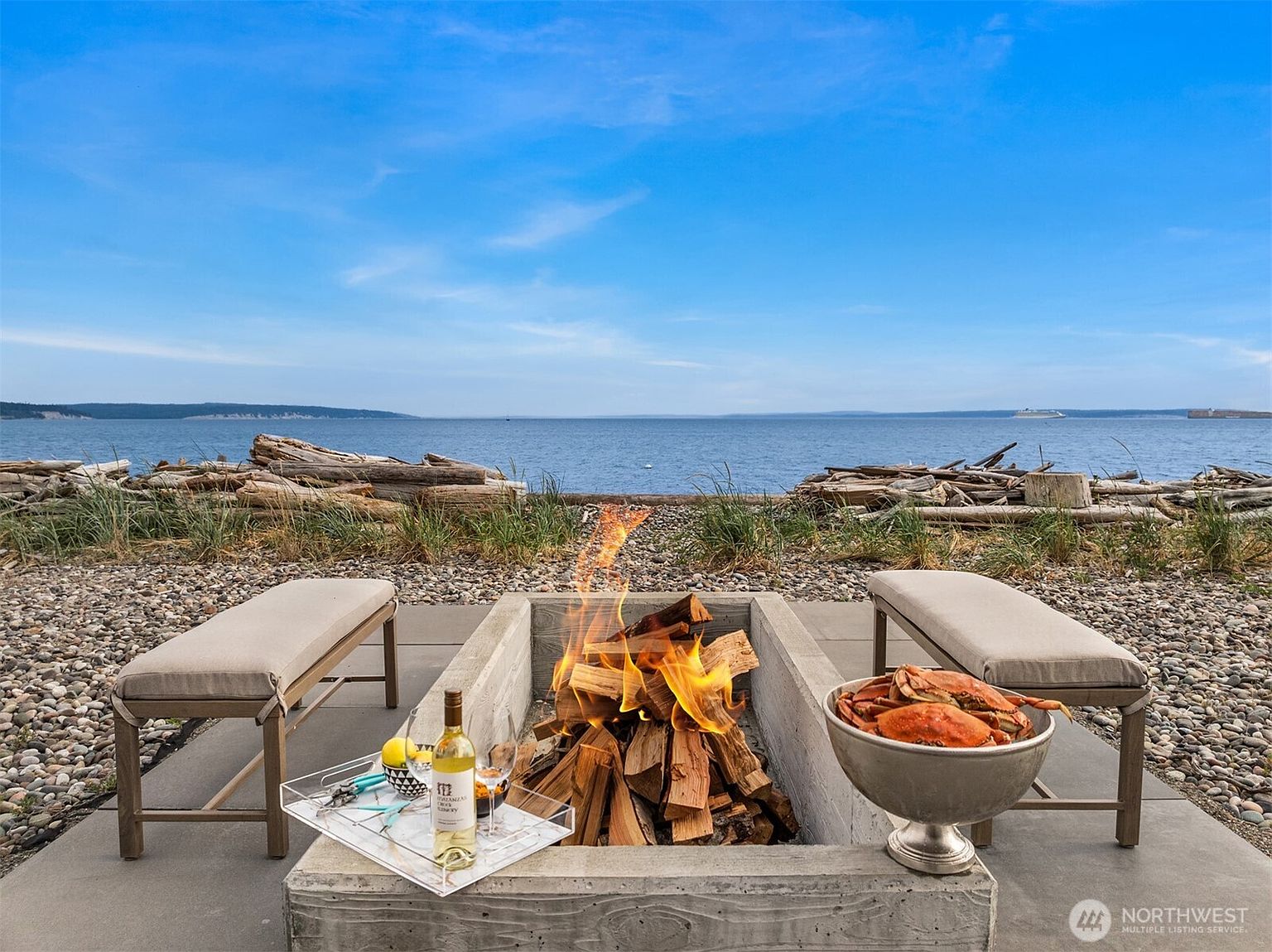This image showcases an inviting outdoor patio area with a stunning waterfront view. A modern concrete fire pit takes center stage, flanked by comfortable benches and a tray with wine and glasses, suggesting relaxation and entertainment. The backdrop features a serene ocean scene with driftwood-strewn shores, creating a picturesque and desirable setting.