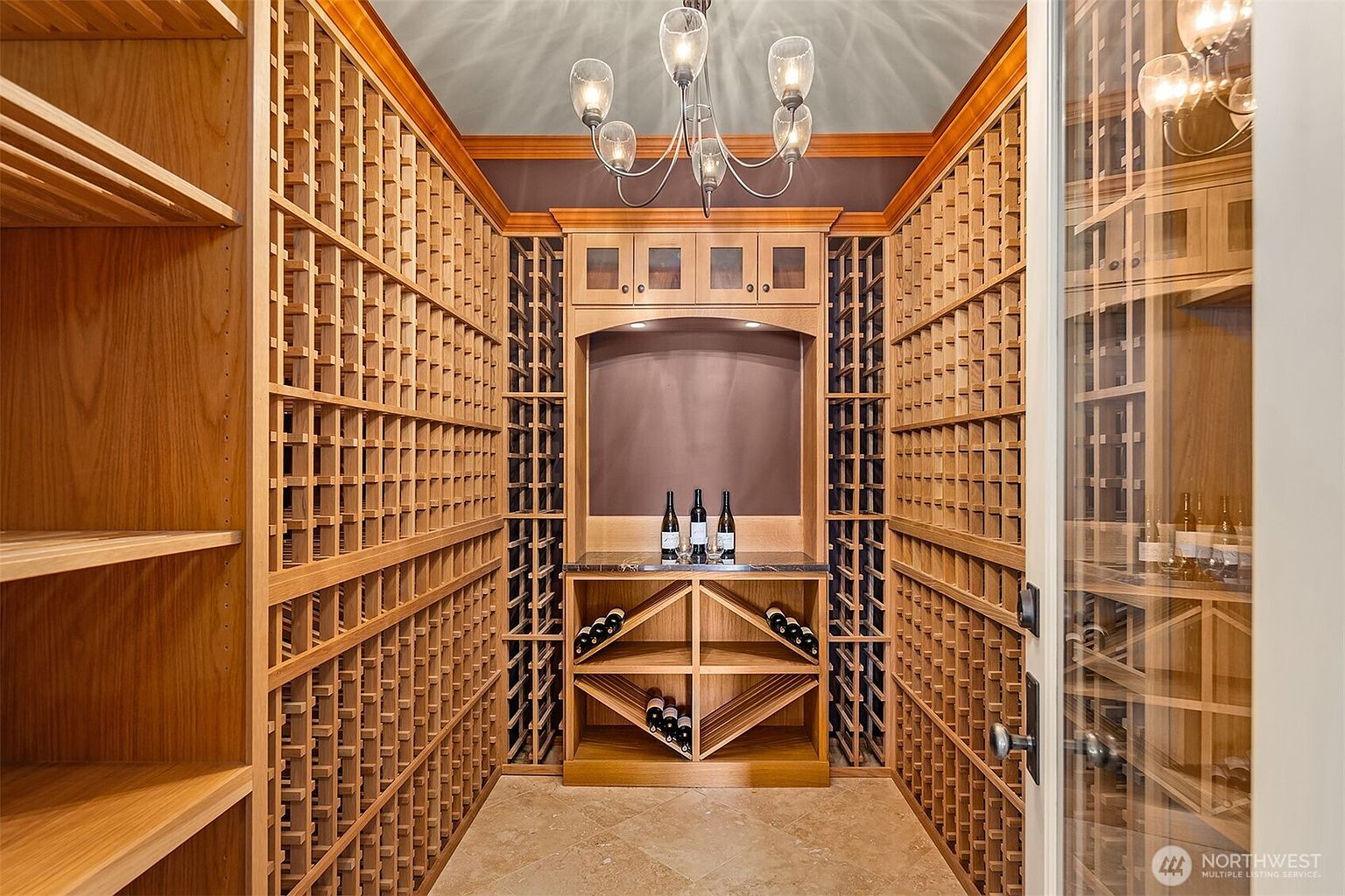 This sophisticated custom wine cellar features extensive floor-to-ceiling wooden racking with dedicated storage for individual bottles and diamond-shaped bins for bulk storage. A central display counter holds three wine bottles beneath a classic chandelier, creating an elegant and inviting atmosphere. The space is framed by rich wood cabinetry and finished with a neutral stone tile floor, offering a refined aesthetic for wine enthusiasts.