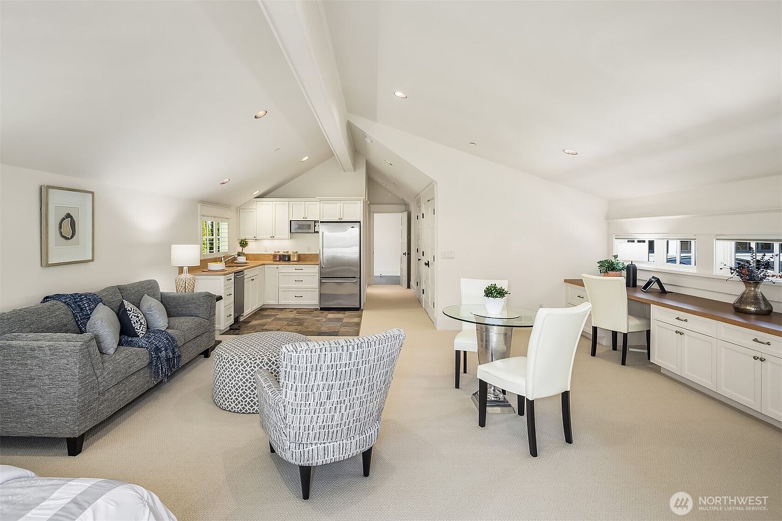 This open-concept living space features a vaulted ceiling, a cozy gray sofa, and a patterned armchair, seamlessly blending into a compact kitchen area and a dedicated workspace. The room is bathed in natural light from multiple windows, creating an airy and inviting atmosphere with a neutral color palette. The perspective is wide-angle, capturing the multi-functional layout that combines relaxation, dining, and productivity in one cohesive area.
