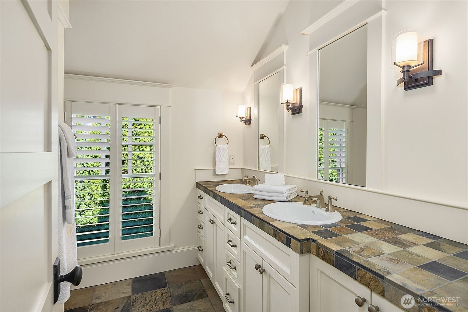 This bathroom features a double vanity with a rustic, multi-colored slate tile countertop and white cabinetry. The space is illuminated by wall-mounted sconces flanking two mirrors, and a set of white plantation shutters allows natural light to filter in. The overall aesthetic combines traditional cabinetry with earthy, textured stone elements for a warm, classic feel.