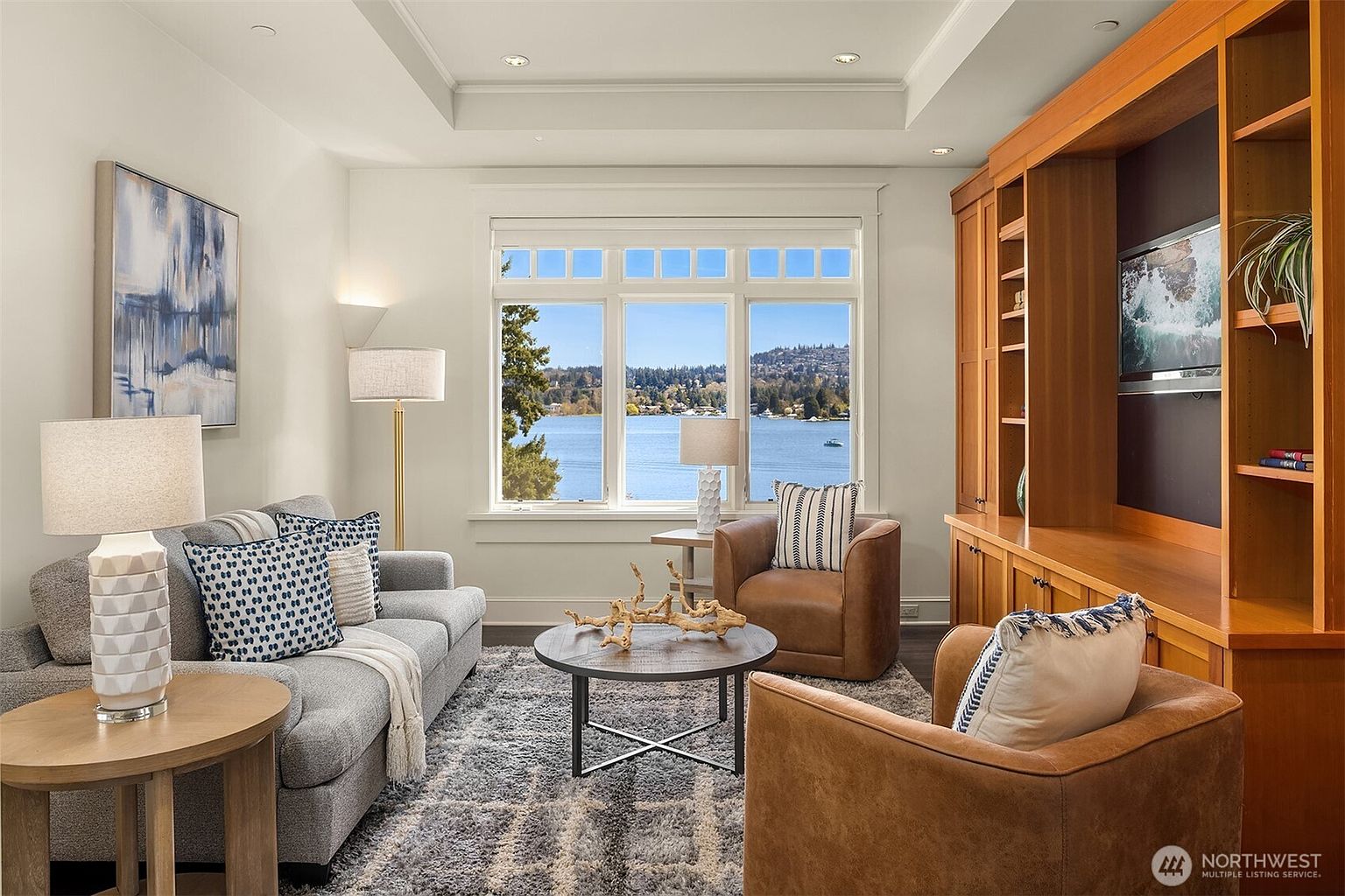 This inviting living room features a cozy gray sofa and two warm leather armchairs arranged around a circular wooden coffee table on a textured area rug. A large built-in wooden entertainment center provides ample storage, while a expansive window offers a stunning, serene view of the lake and surrounding trees. The room is accented with soft lighting from floor and table lamps, creating a comfortable and sophisticated atmosphere.